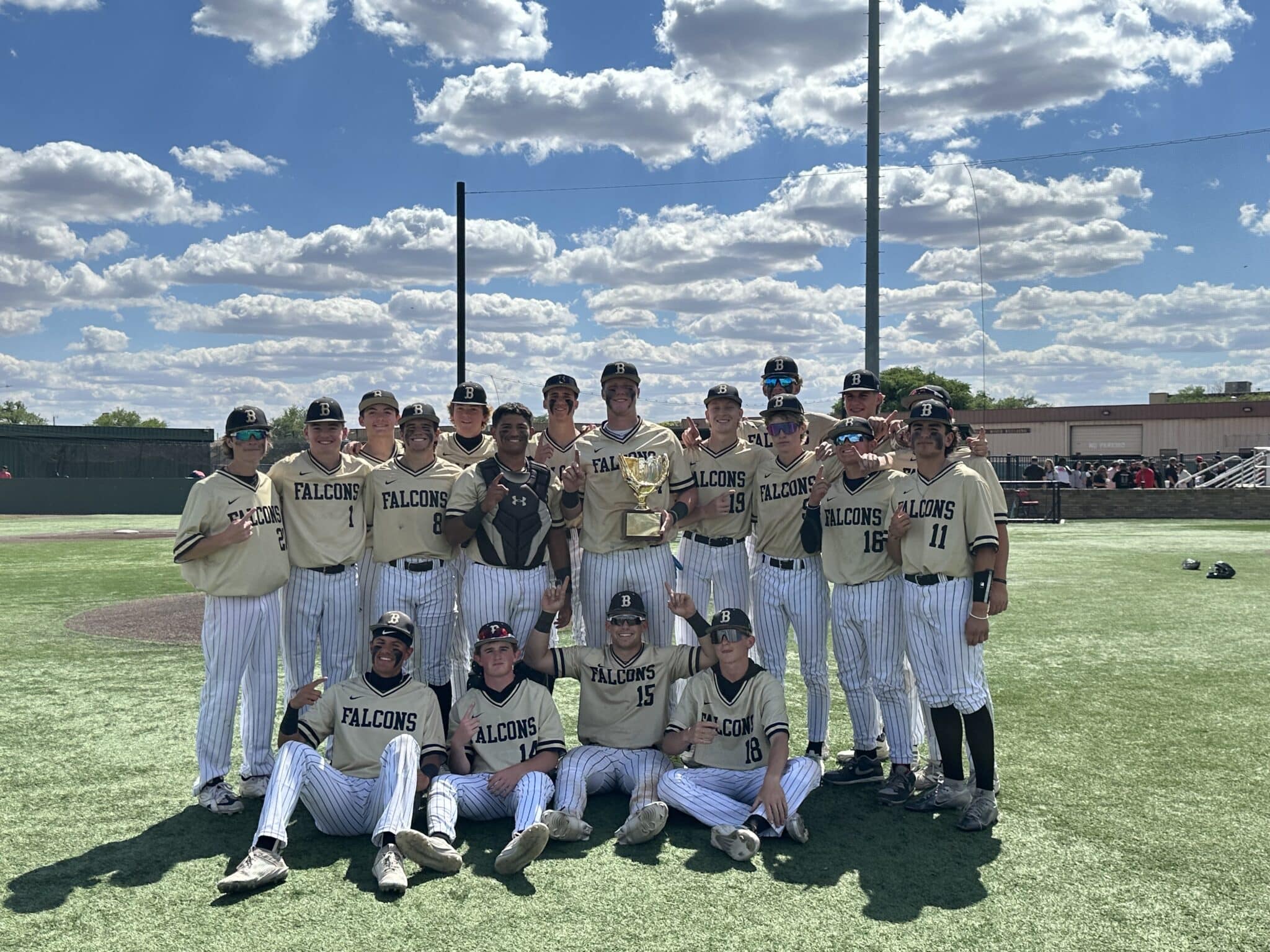 Falcons baseball team posing together on the field with a trophy after a win, sunny sky above.