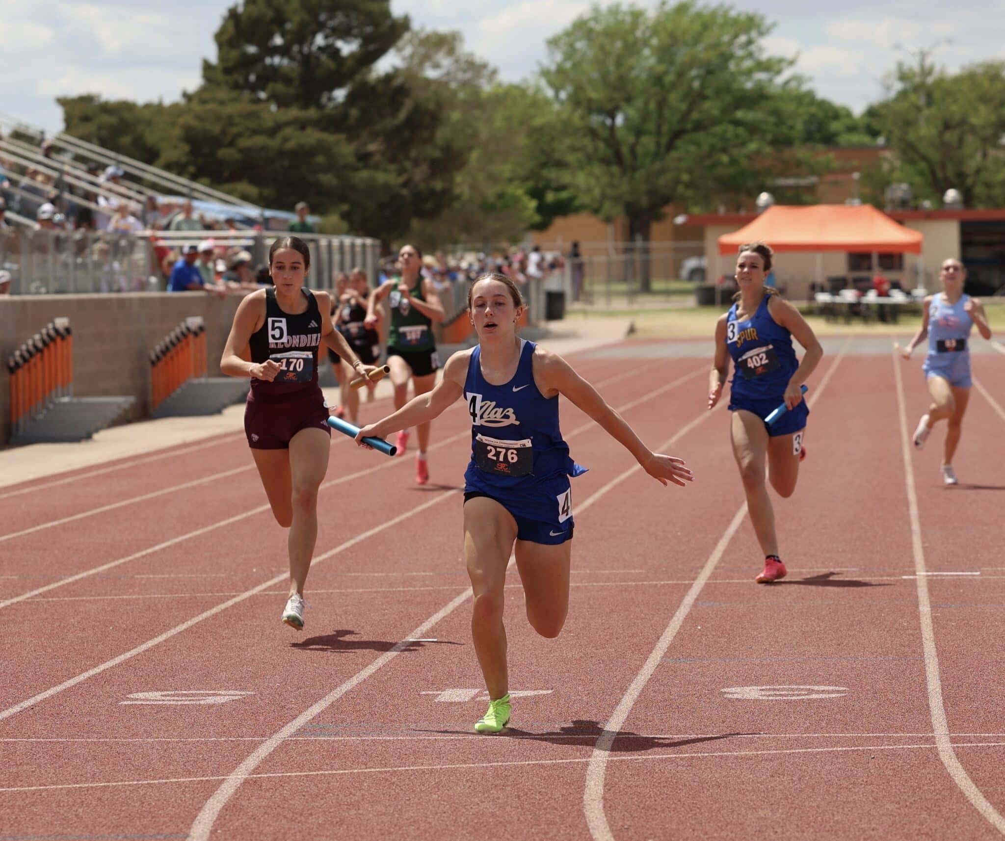 Female sprinter in blue leads a track race on a red running track while competitors chase behind.