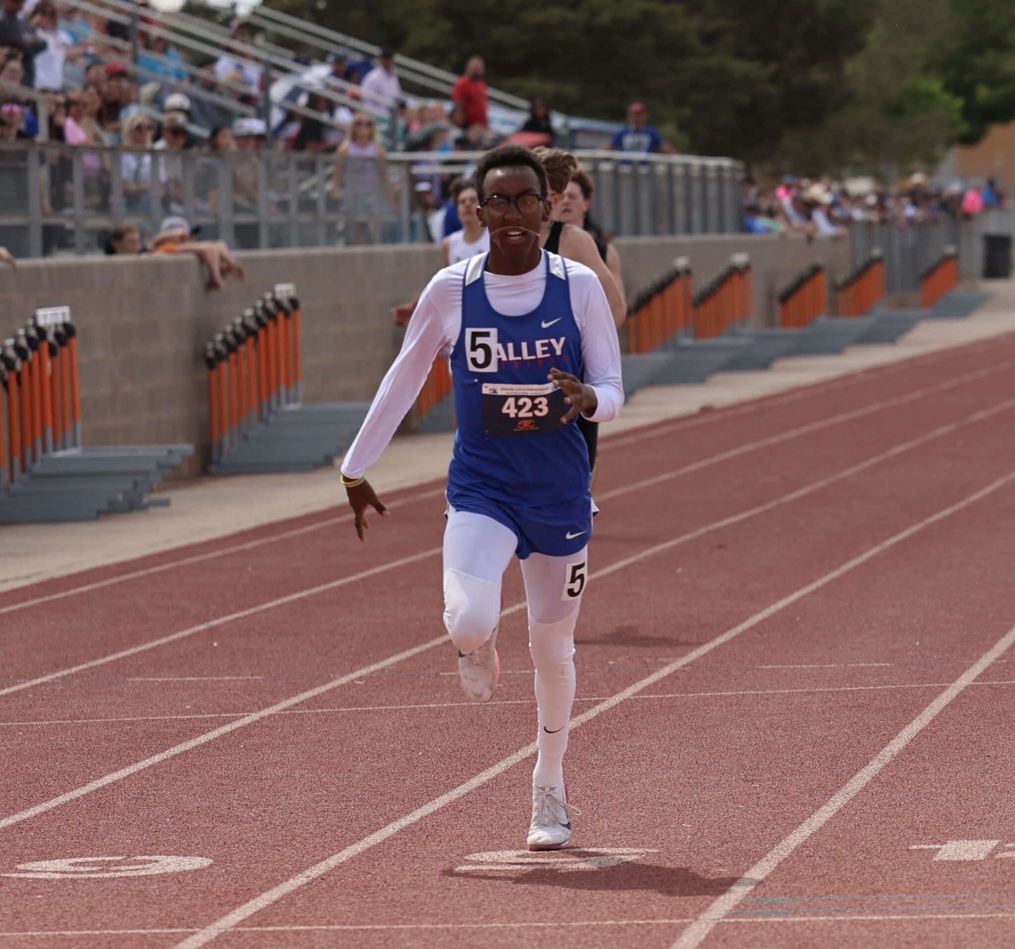 Male sprinter in a blue and white uniform with bib number 423, running on a red track while spectators watch from the stands.