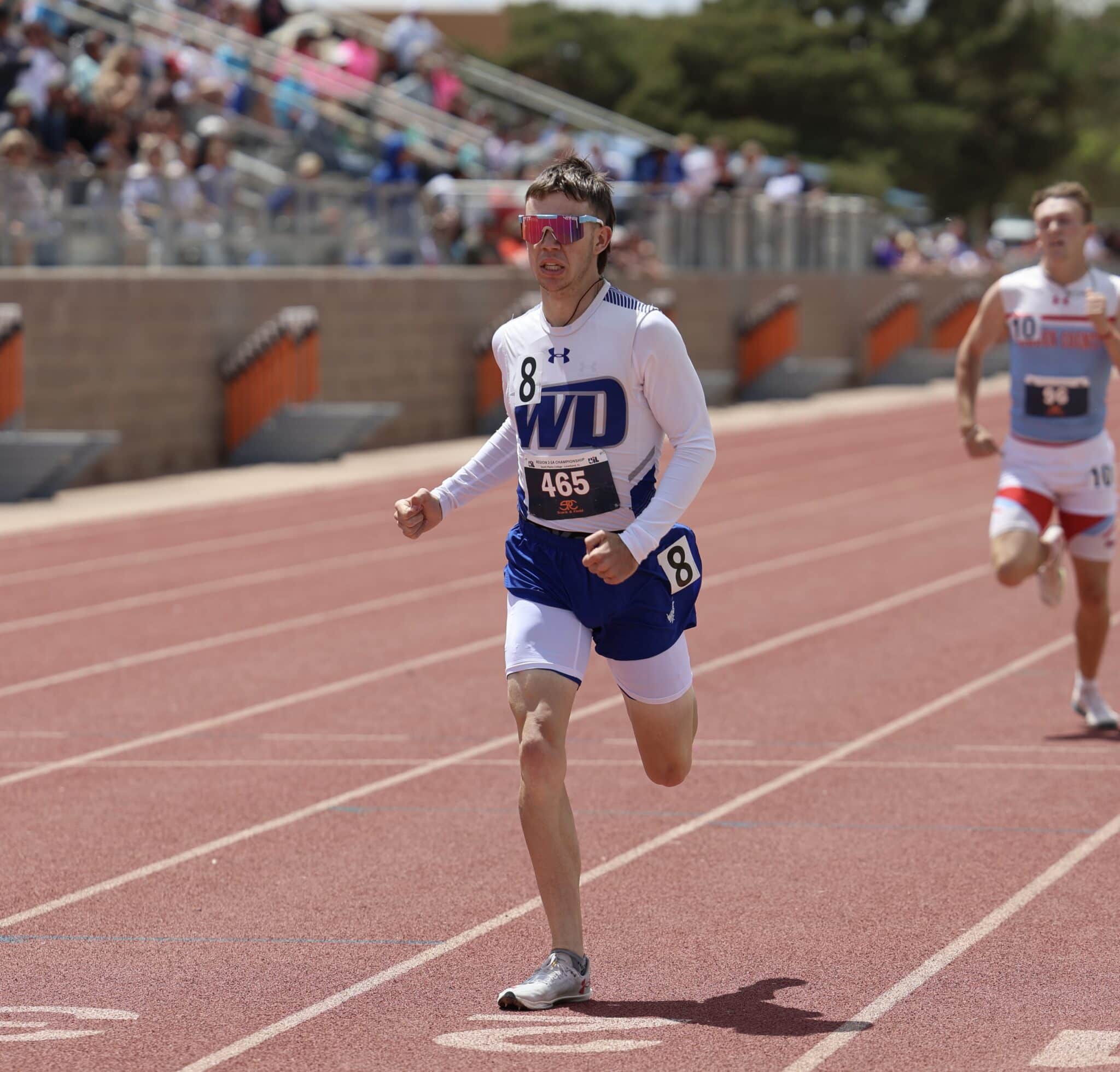Male runner in a white and blue uniform with pink mirrored sunglasses, sprinting on a red track as blurred spectators watch from the stands.