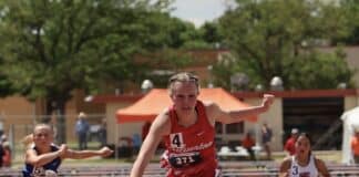 Female athlete in red clears a hurdle during a middle-distance track race, with other runners behind on the track.