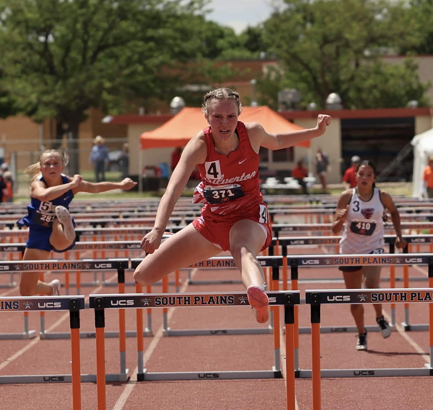 Female athlete in red clears a hurdle during a middle-distance track race, with other runners behind on the track.