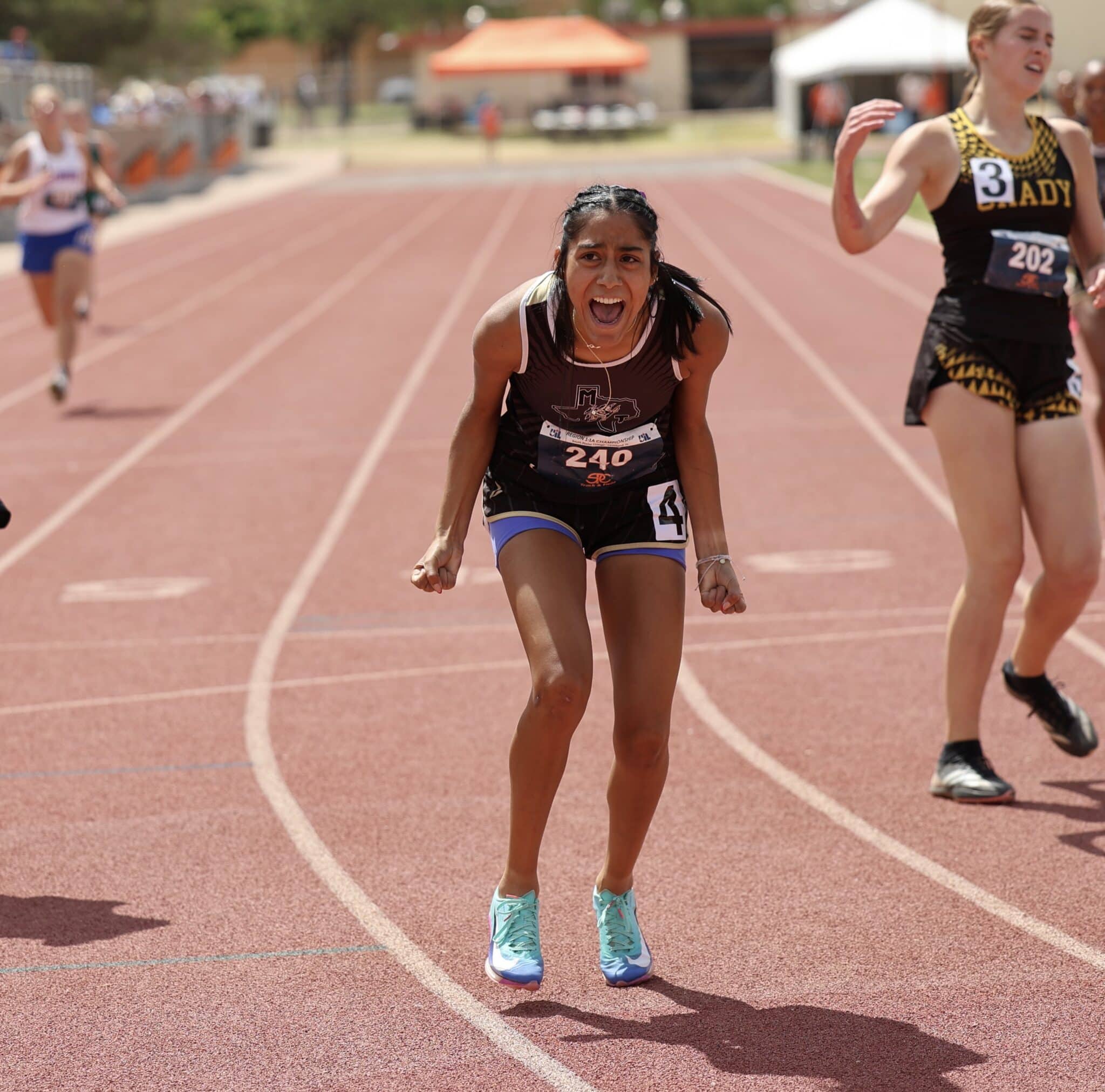 Female sprinter in black and white uniform with bib 240 running toward camera on red track; other runners blurred in background.