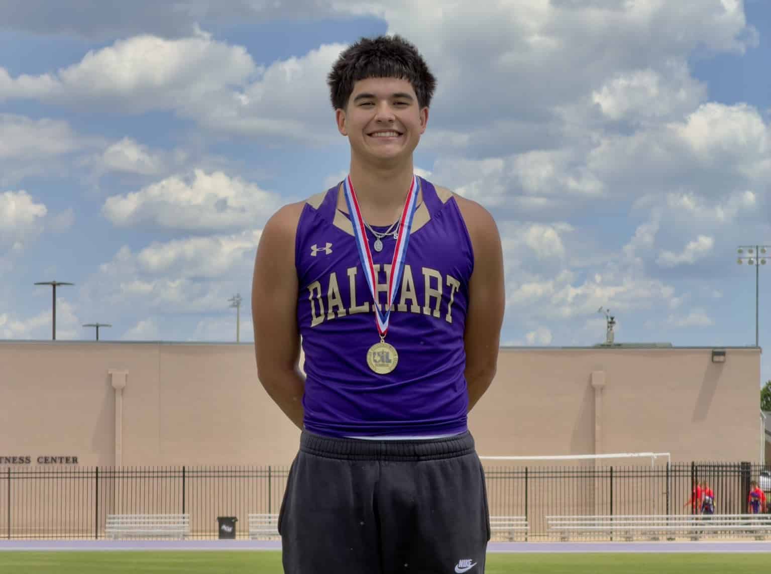 Male student-athlete in a purple Dalhart jersey with a medal around his neck, smiling on a track field against a blue sky with clouds.