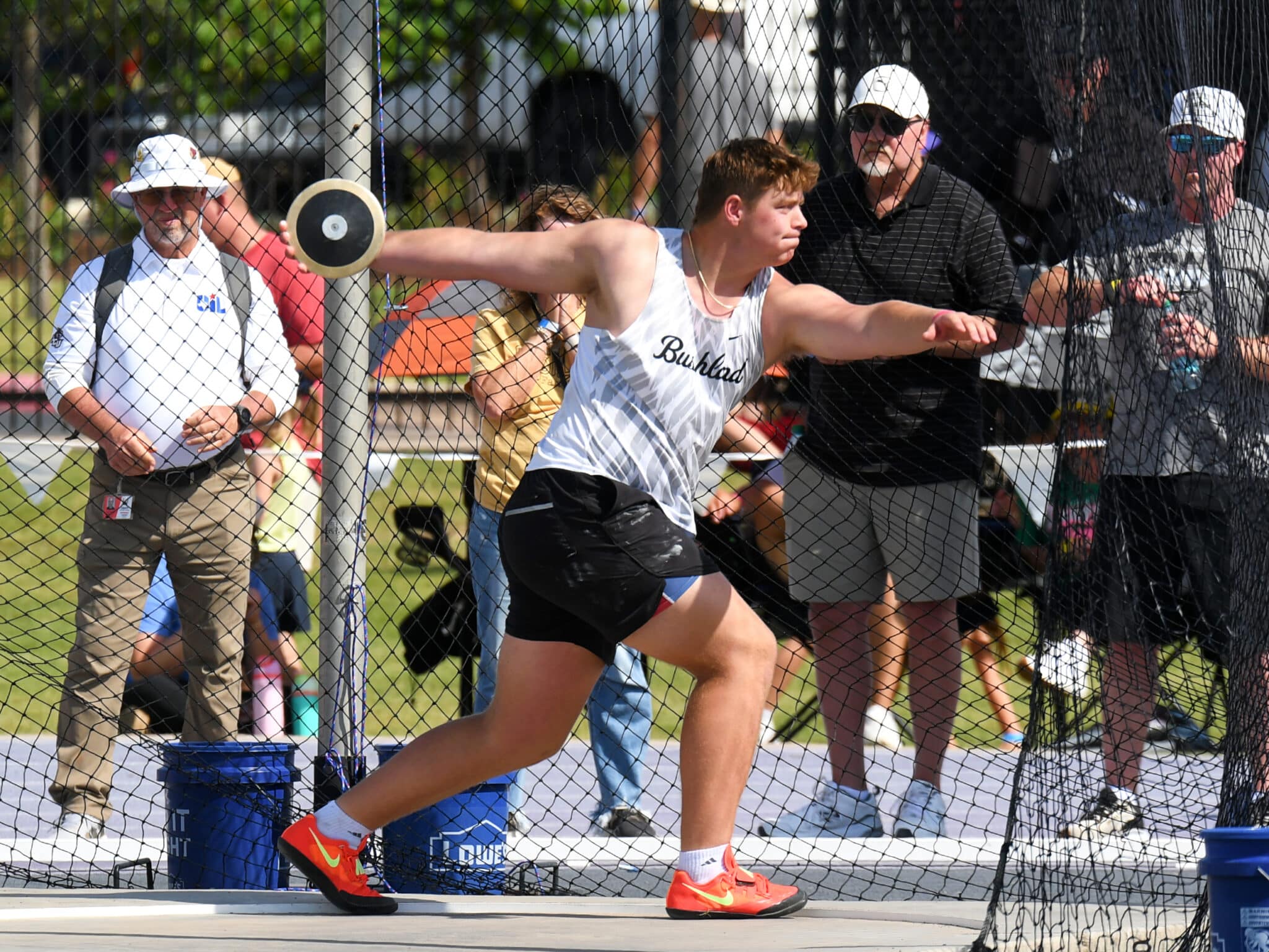 Athlete in a white sleeveless shirt throws a discus through a safety net, with spectators watching nearby.