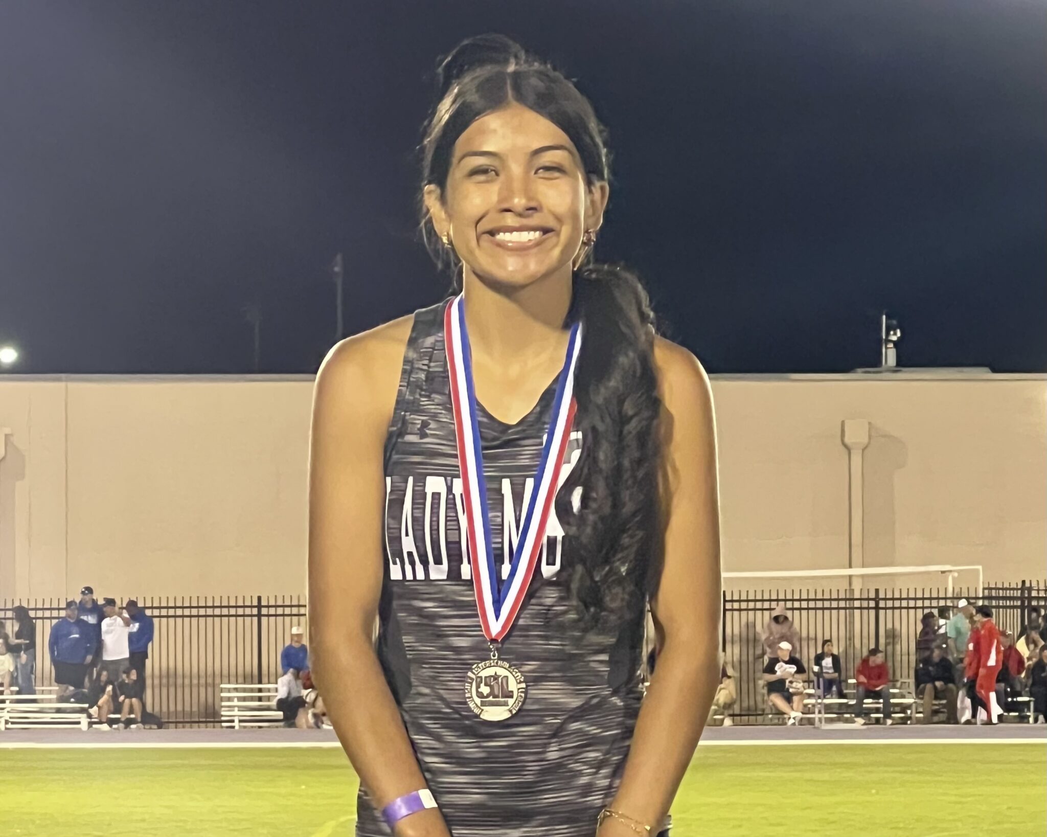 Smiling young female athlete with medals around her neck, posing after a nighttime track event at the stadium.