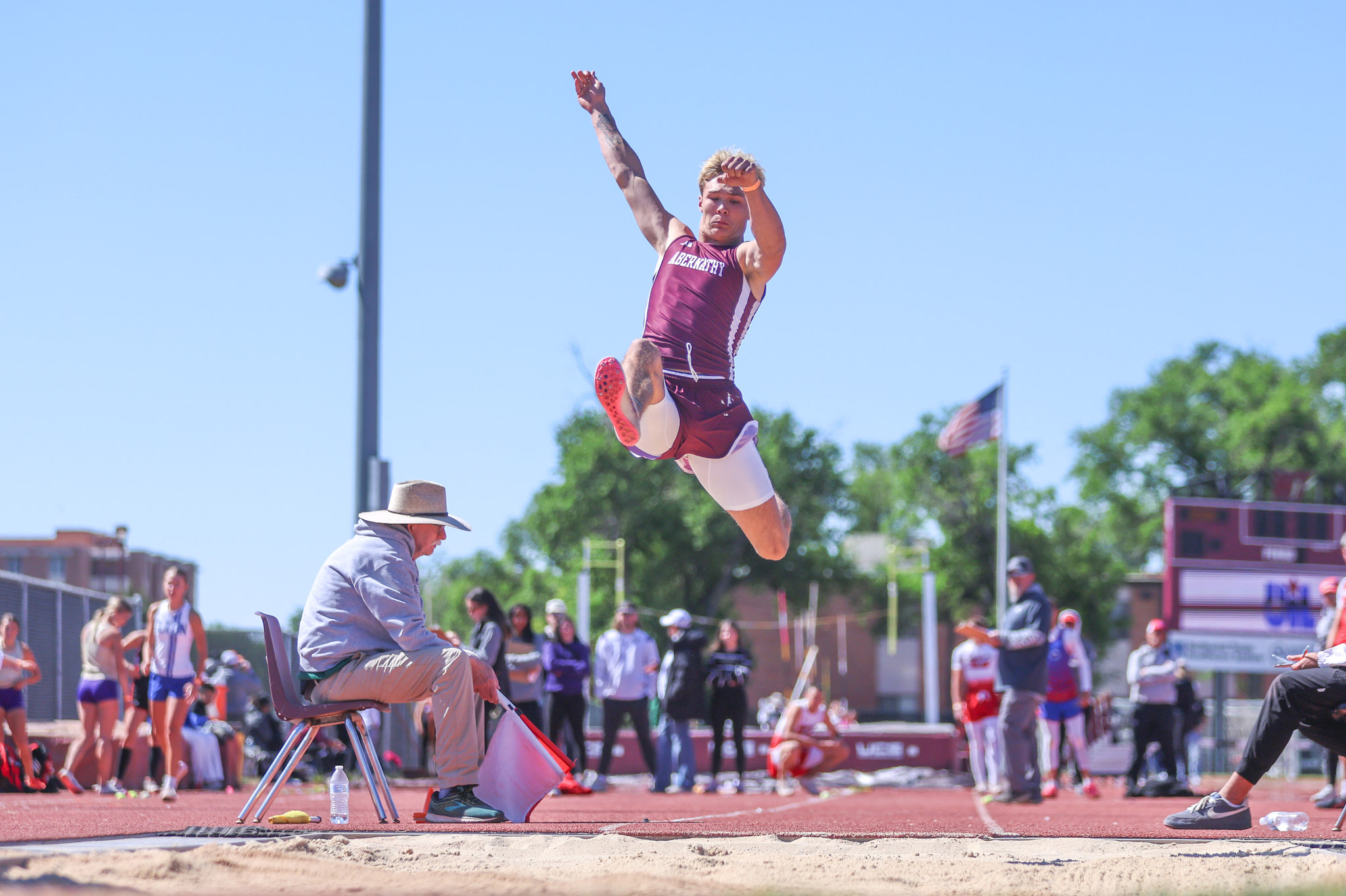 Male athlete in a maroon uniform mid-jump during a sunny track meet, leaping toward a sand pit with spectators and officials watching in the background.