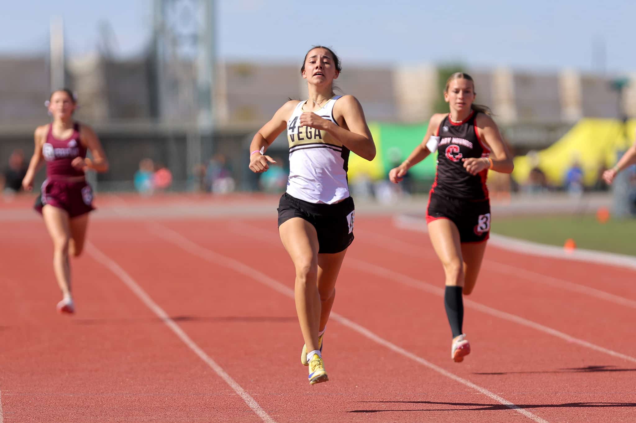 Three female sprinters race on a red outdoor track, with the central runner in a white top leading the pack.