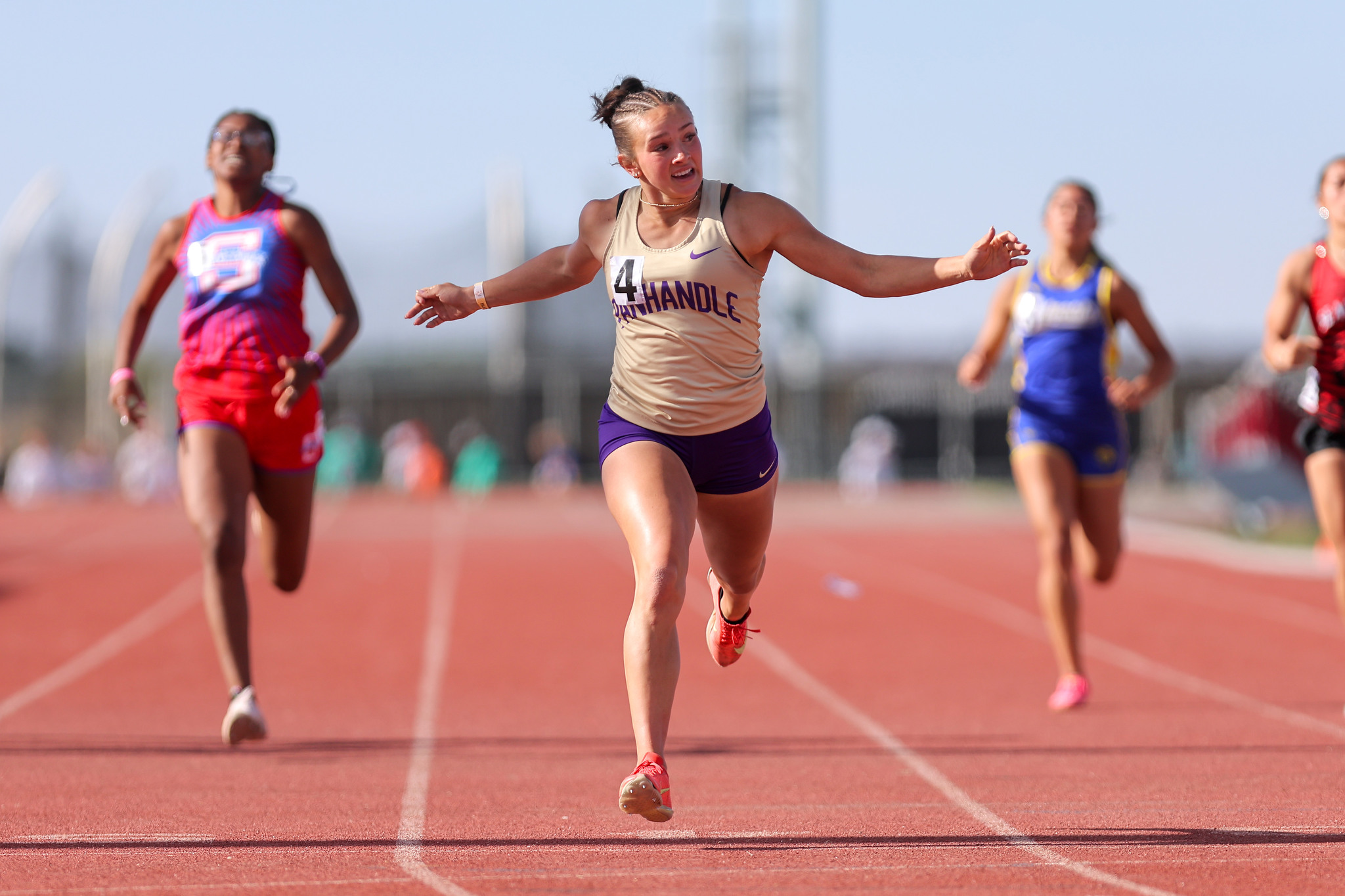 Female track sprinter in a beige top and purple shorts crosses the finish line with arms outstretched on a red track, other competitors blurred in the background on a sunny day.
