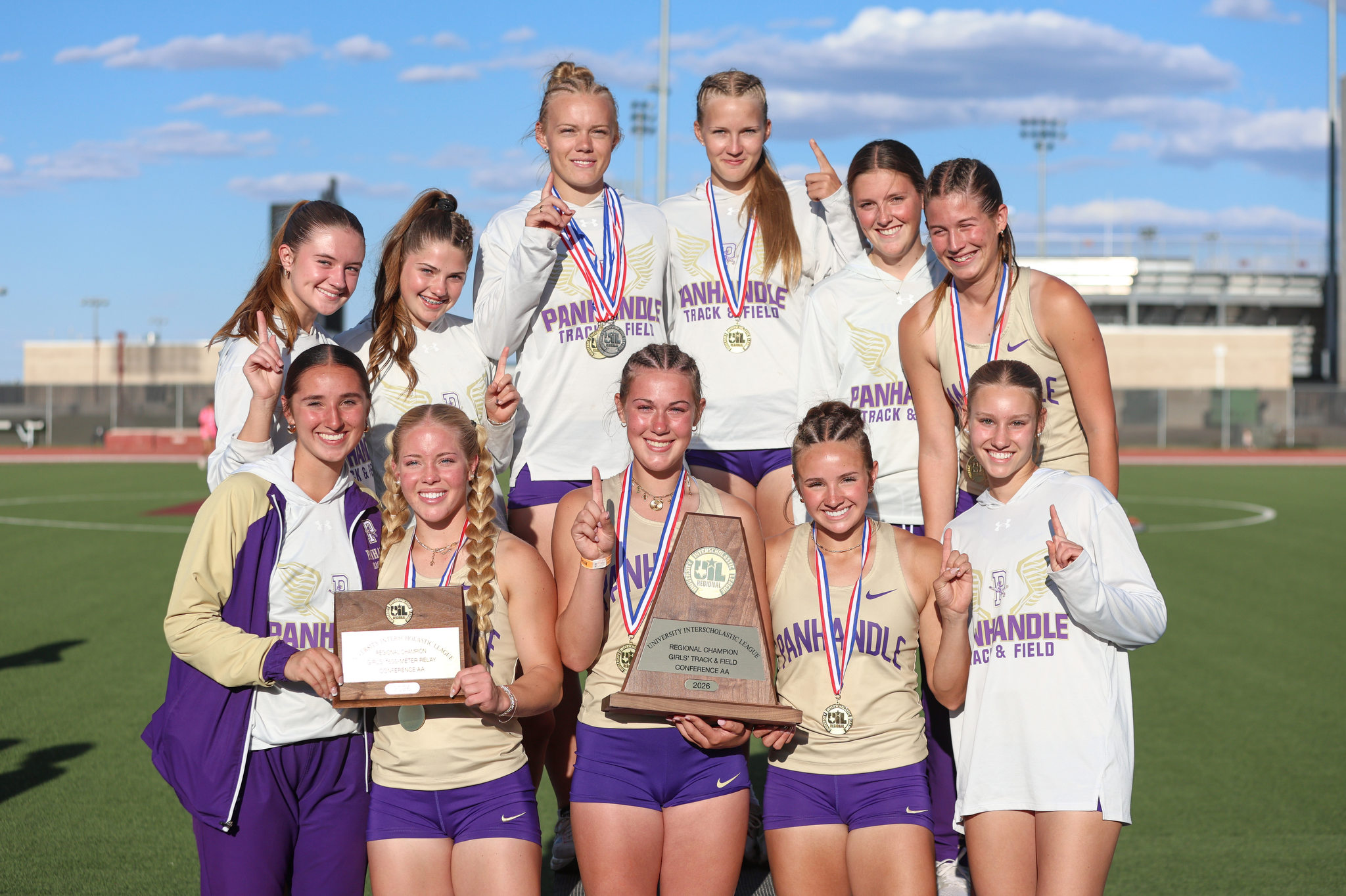 Group of female track athletes posing on a field, wearing matching uniforms and medals after a win, holding trophies.