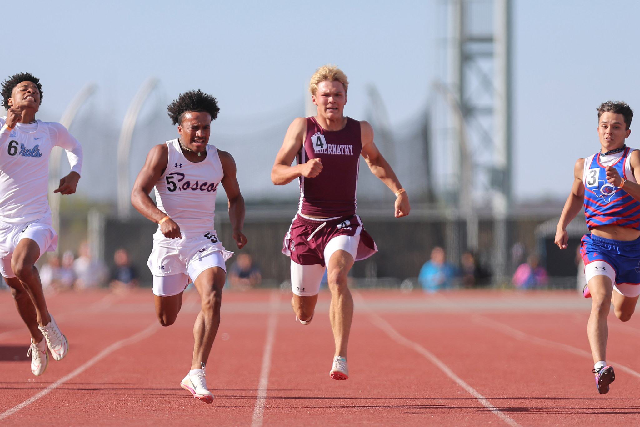 Four male sprinters race down a red track, bibs 6, 5, 4, and 3 visible, with the lead runner in maroon at center.