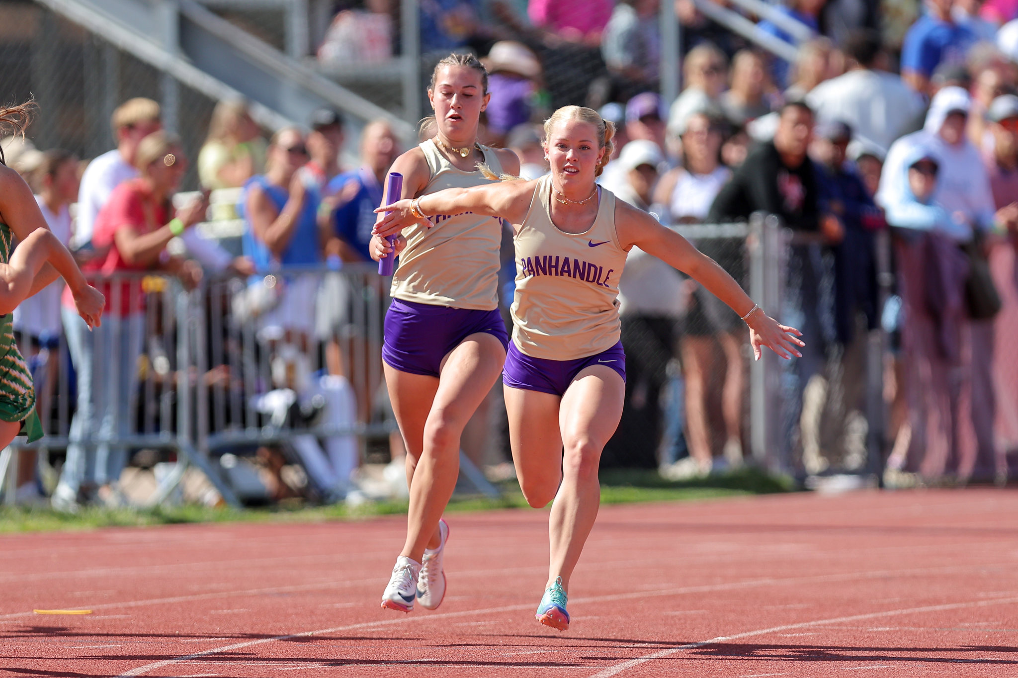 Two female relay runners in beige tops and purple shorts sprint on a track, one passing a purple baton while spectators watch from behind fencing.
