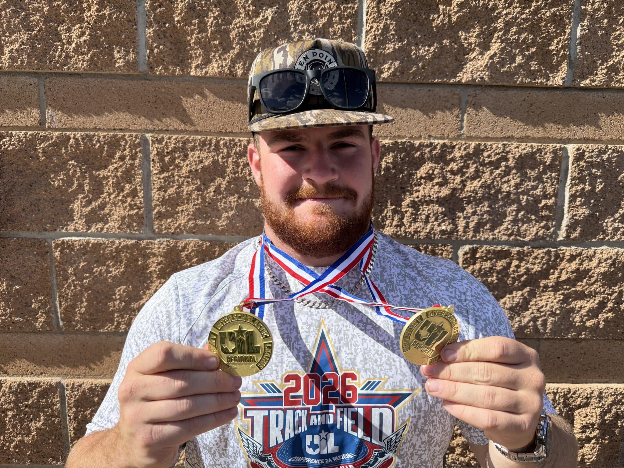 Man posing against a brick wall, wearing a camo cap with sunglasses on top and multiple gold medals around his neck.