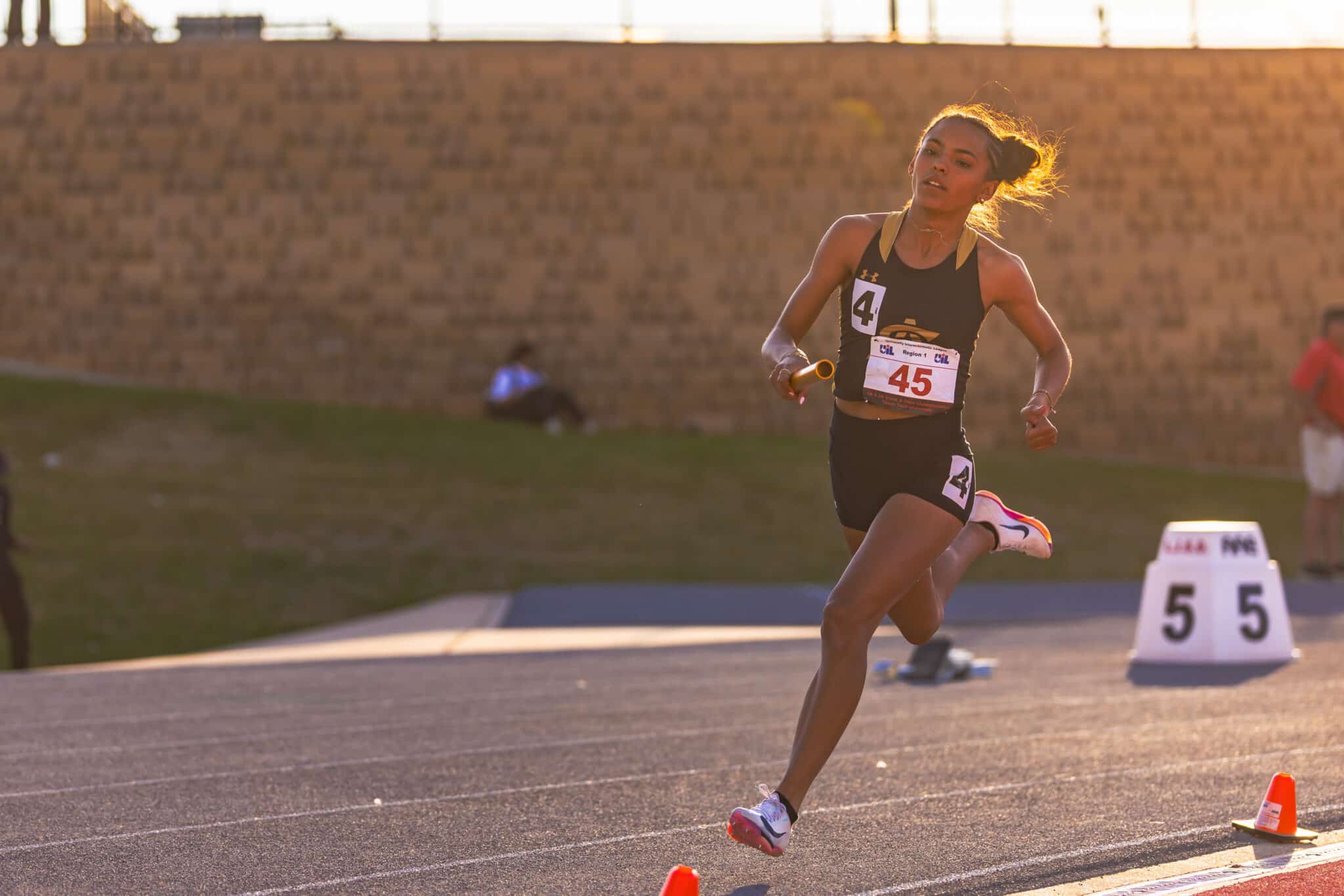 Female sprinter wearing bib 45 running on a track with orange cones nearby, sunset lighting in the background.