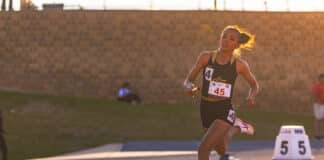 Female sprinter wearing bib 45 running on a track with orange cones nearby, sunset lighting in the background.