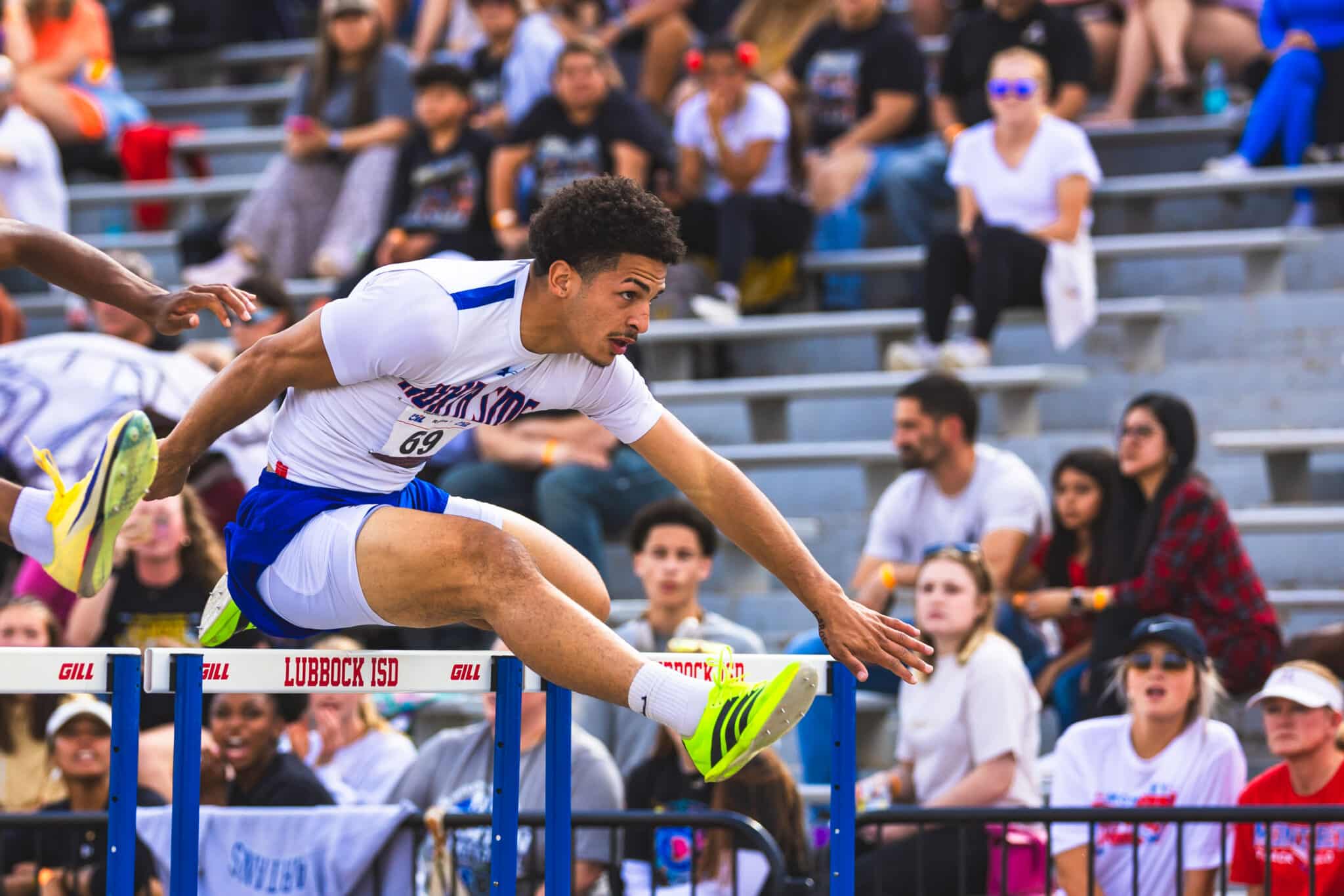 Male athlete in white and blue uniform leaps over a hurdle during a track race, neon green shoes visible, crowd in bleachers behind him.