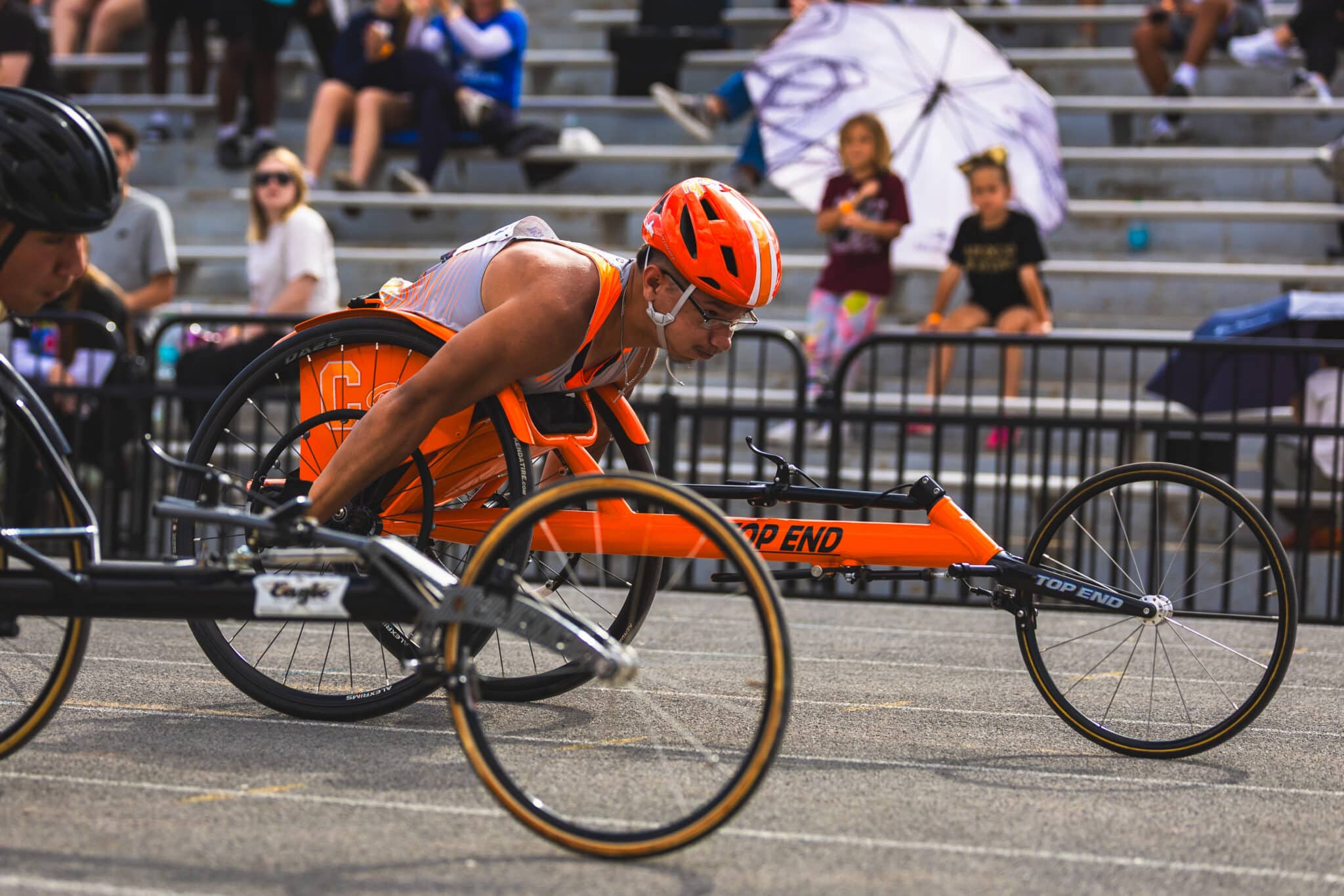 Wheelchair racer in an orange racing chair and helmet leans forward during a race, with spectators in bleachers behind a barrier.