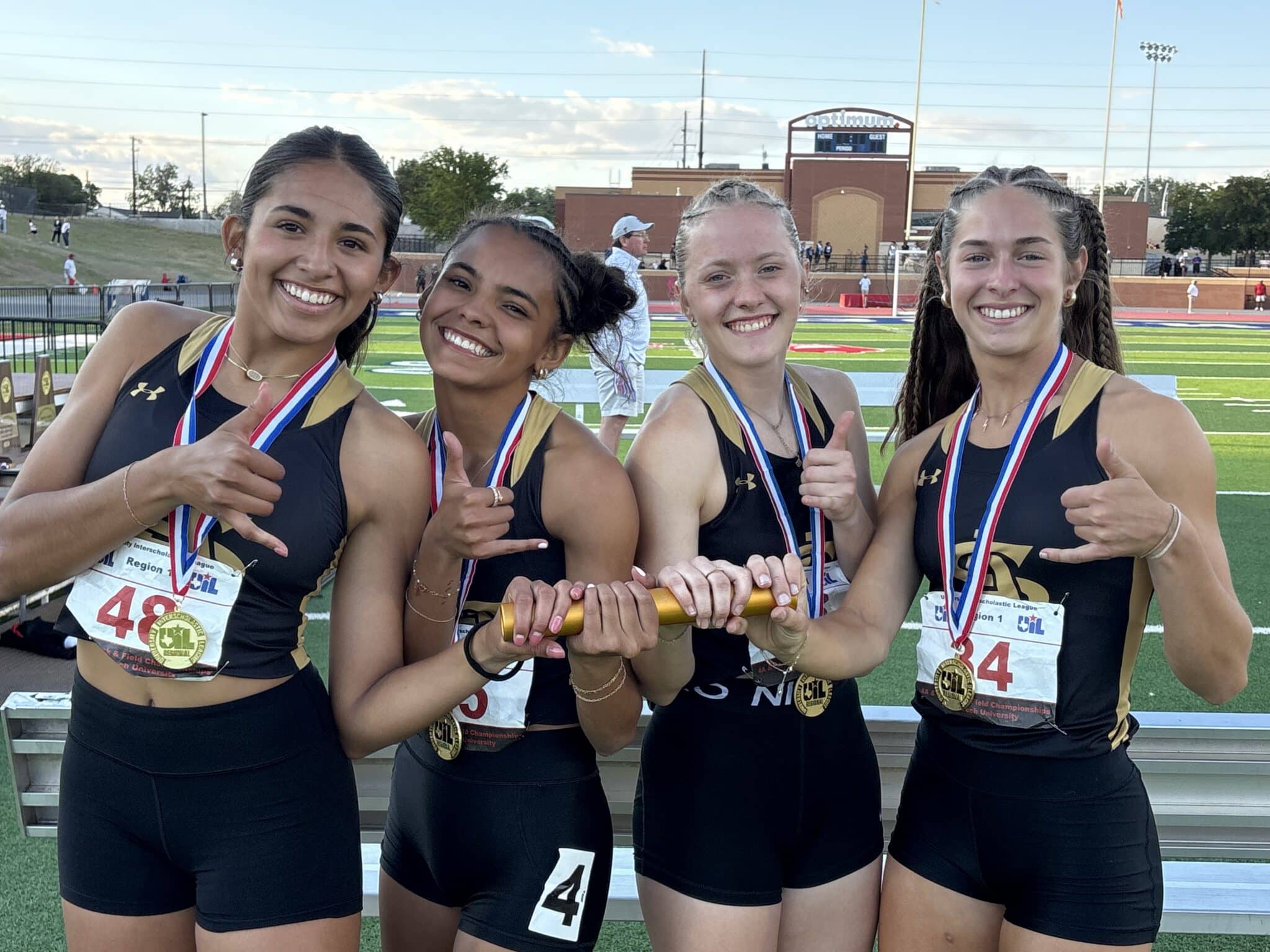 Four young female runners in black-and-gold uniforms smile and thumb-up after a race, medals around their necks on a track field. One holds a yellow baton; bib numbers visible in front. Stadium and scoreboard in background.