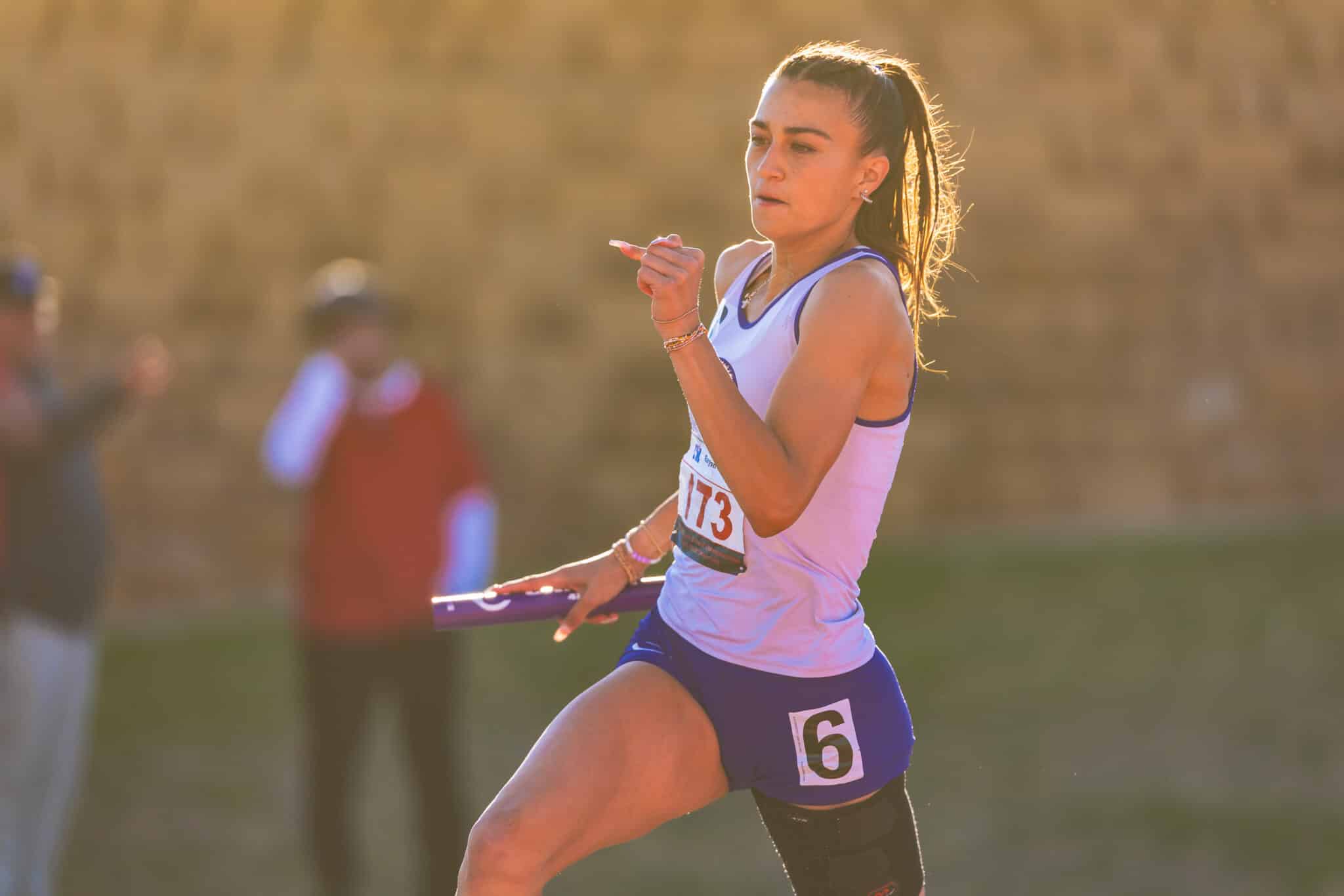 Female sprinter in lavender during a relay race, holding a baton and running forward in a sunlit field.