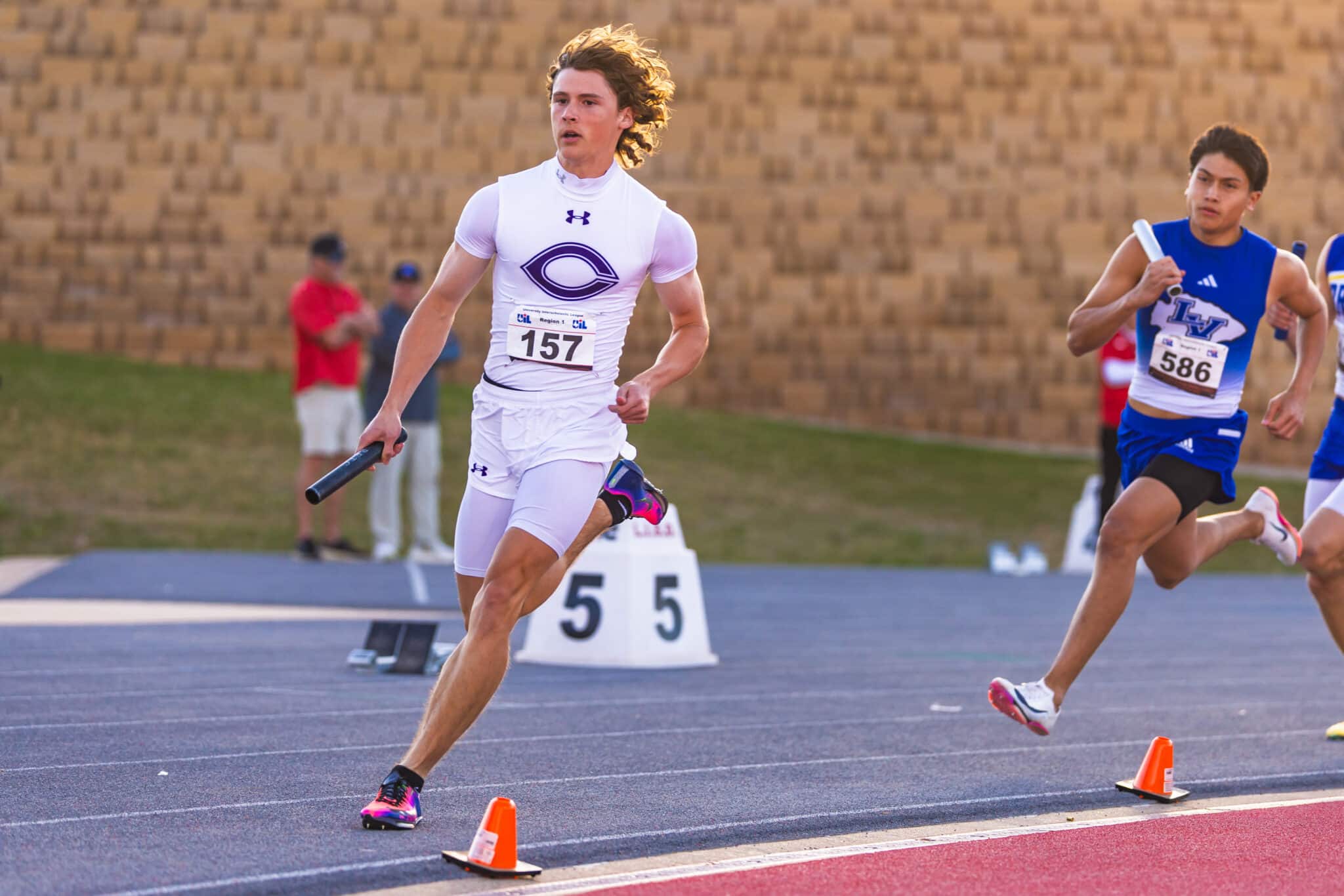 Young male sprinter in a white uniform with bib 157 leading a relay-style race on an outdoor track, orange cones along the lane.