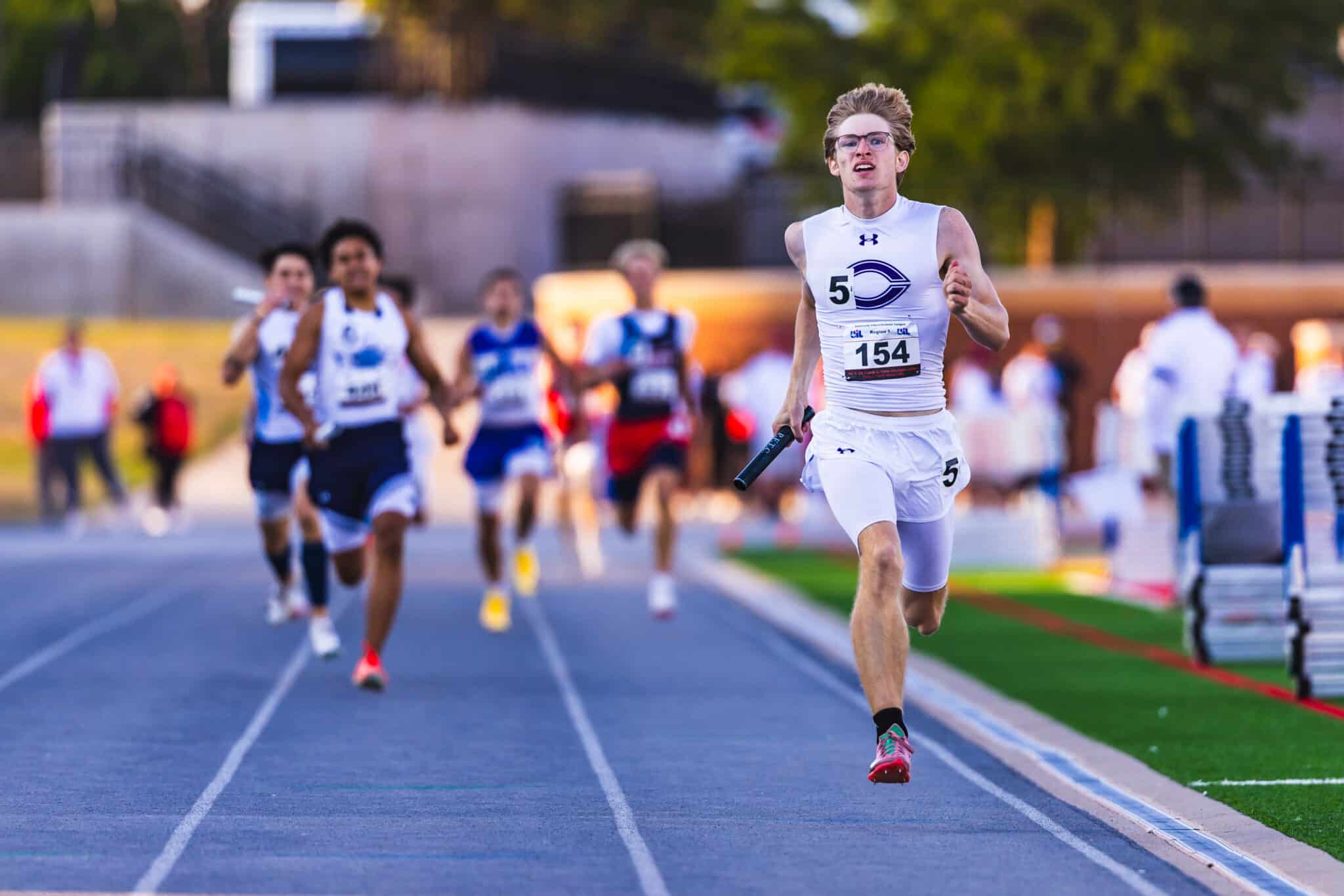 Male sprinter in white uniform leading a relay race on an outdoor track, holding a baton as teammates chase behind him.