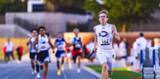 Male sprinter in white uniform leading a relay race on an outdoor track, holding a baton as teammates chase behind him.