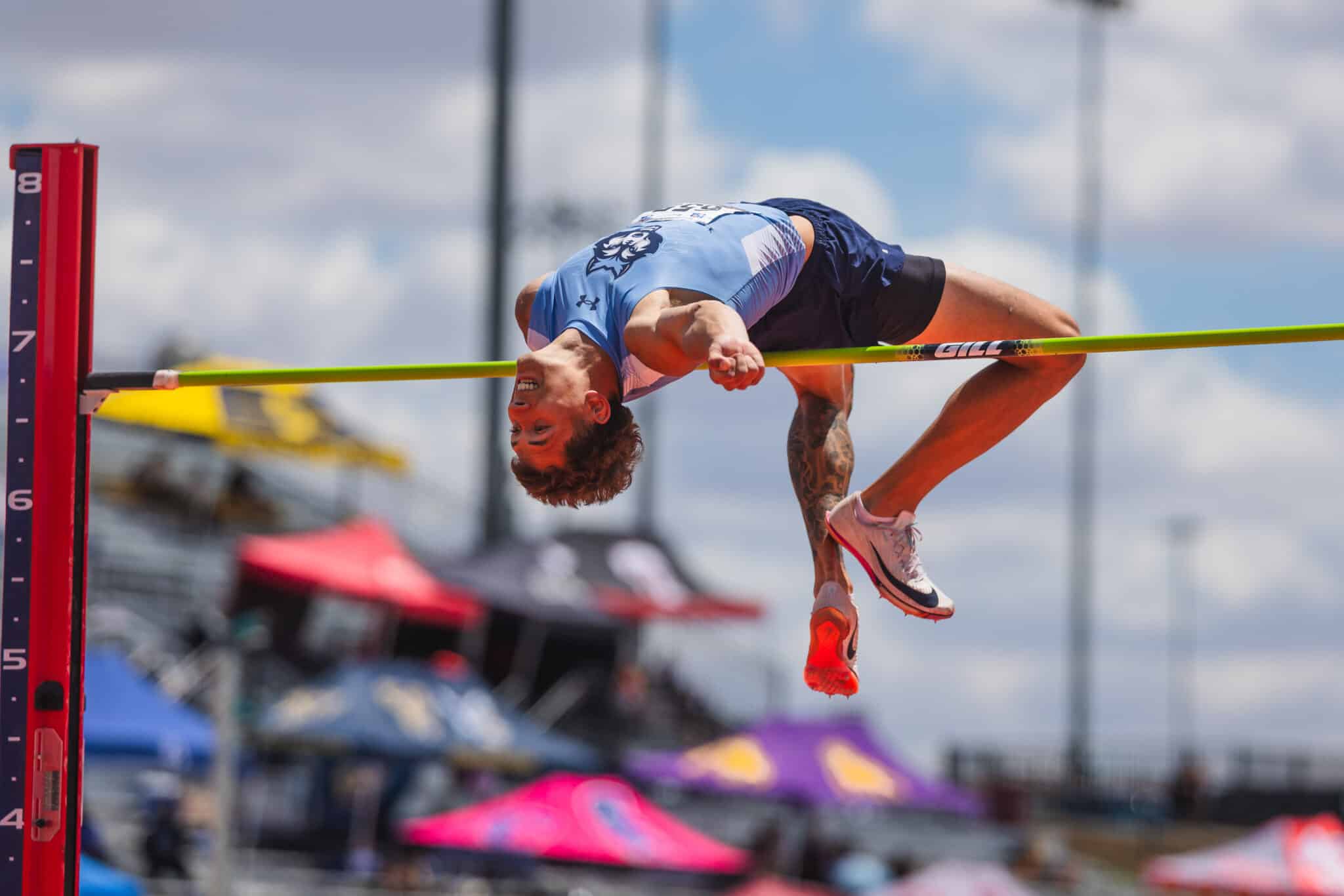 Athlete in a light-blue uniform clears a bright lime-green high jump bar during an outdoor track meet, red support on the left.