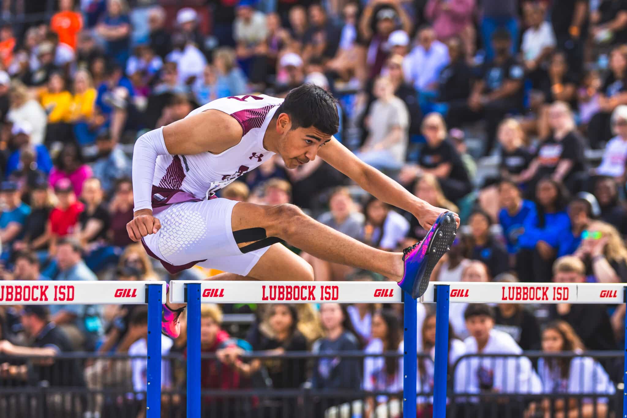 Male hurdler in white maroon uniform clears a hurdle during a crowded track meet; vibrant banners read LUBBOCK ISD.