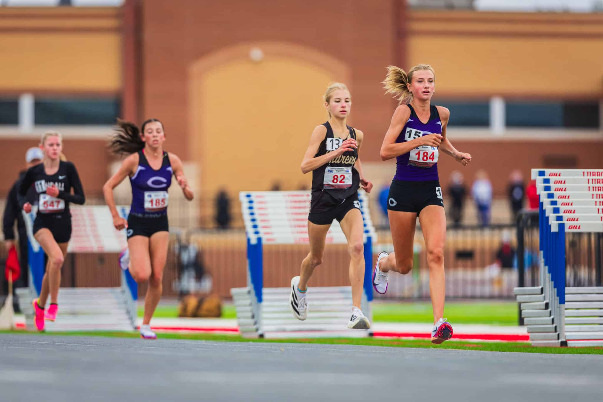 Female athletes compete in a hurdles race on an outdoor track, bib numbers visible (184 front, others 82, 13).