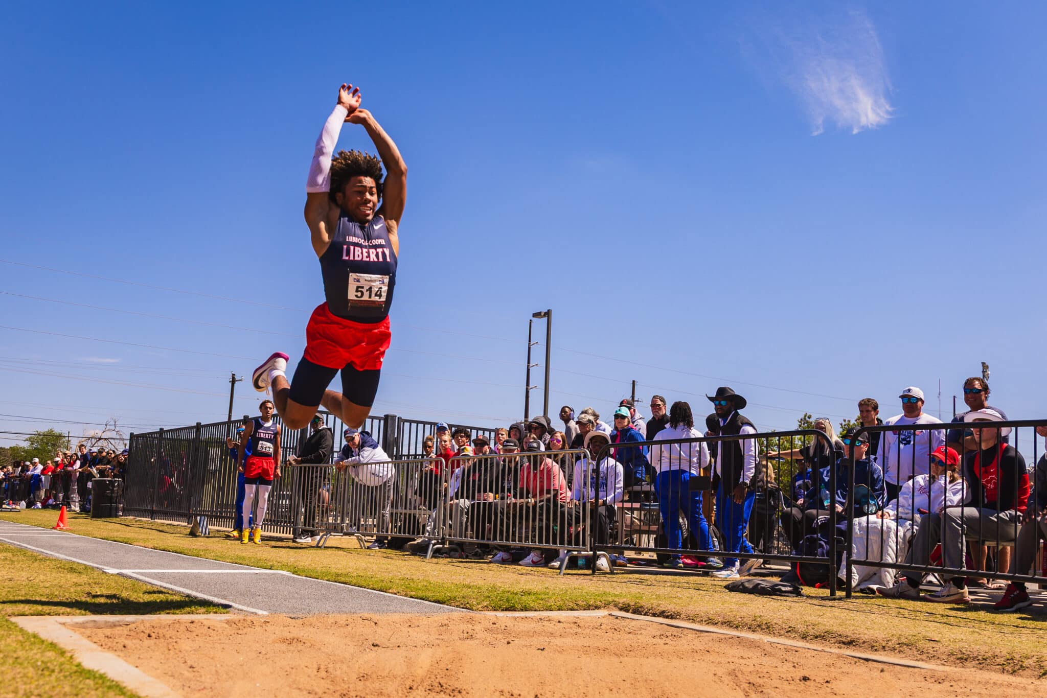 Athlete in a navy singlet with 'LIBERTY' jumps mid-air during a race, bib 514, with spectators behind a barrier against a blue sky.