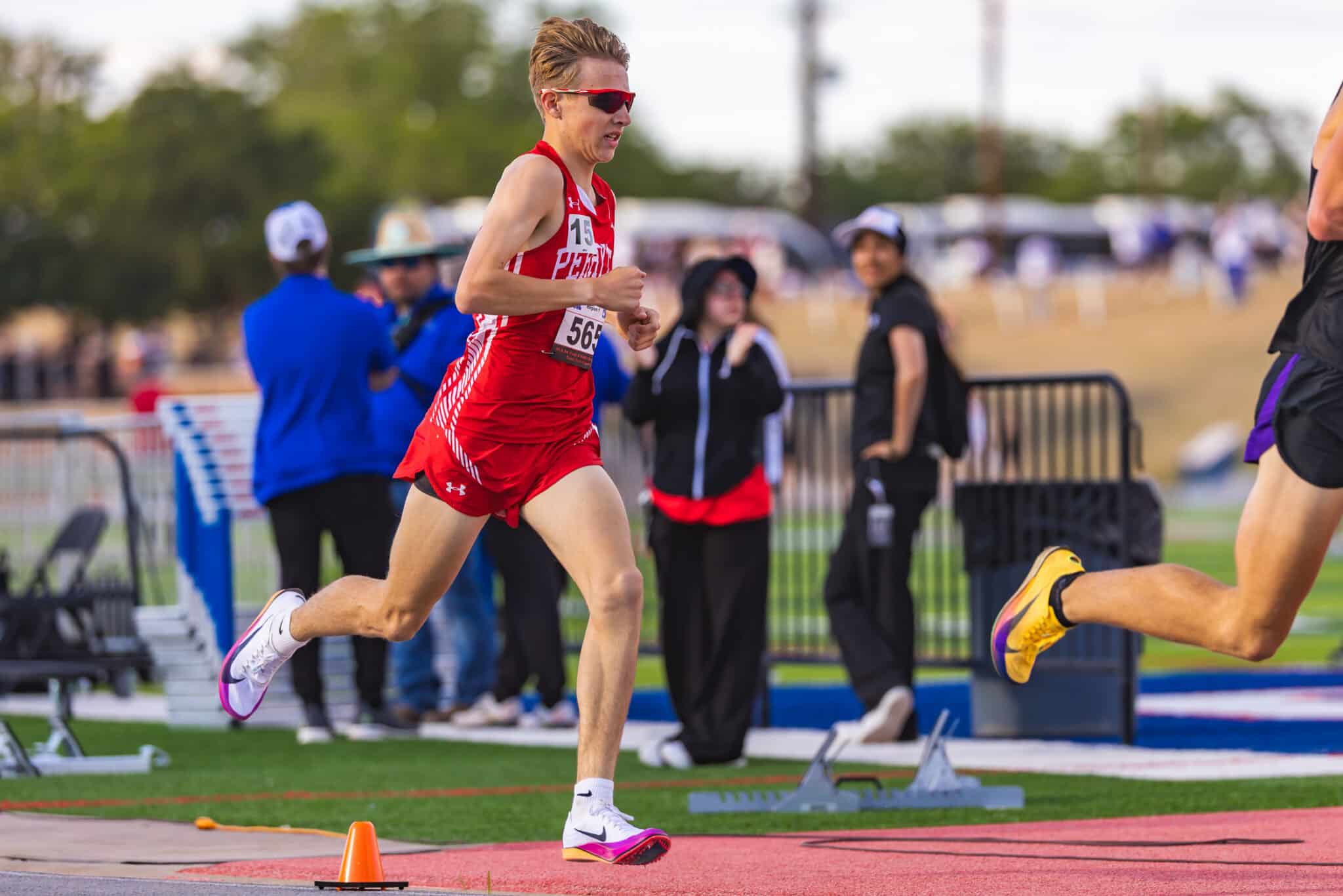 Male runner in red uniform sprinting on a track, bib number 56, with spectators and equipment in the blurred background.
