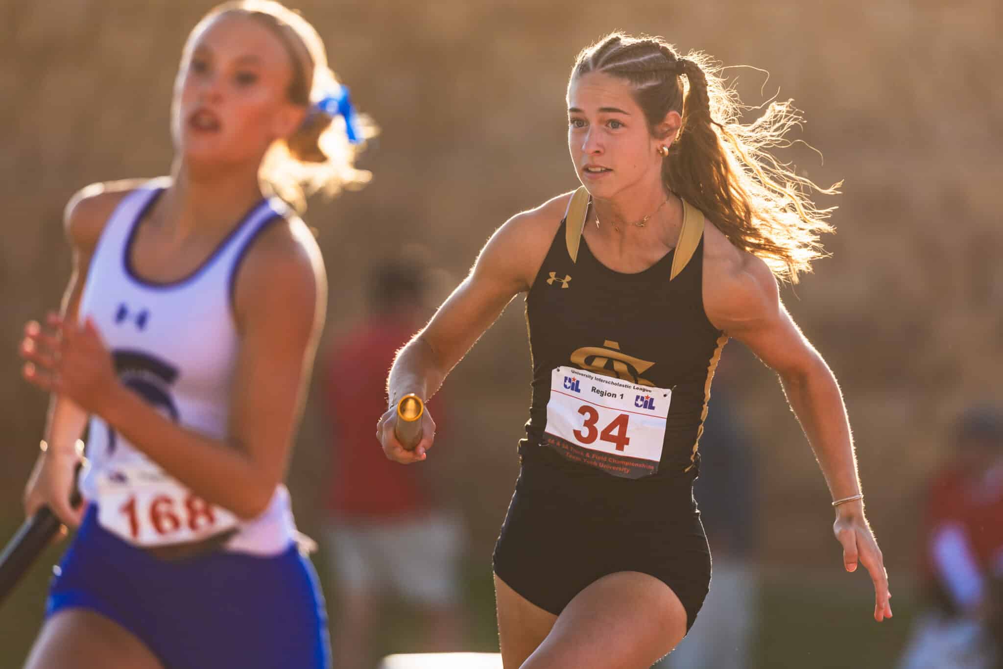Two female sprinters relay in progress: foreground runner in black with bib 34 holding a baton, sunlit backdrop.