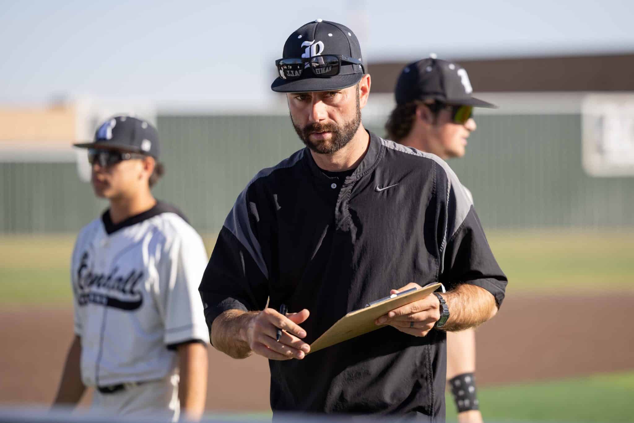 Baseball coach in a black uniform holds a clipboard and notes while two players practice in the background on a sunny field.