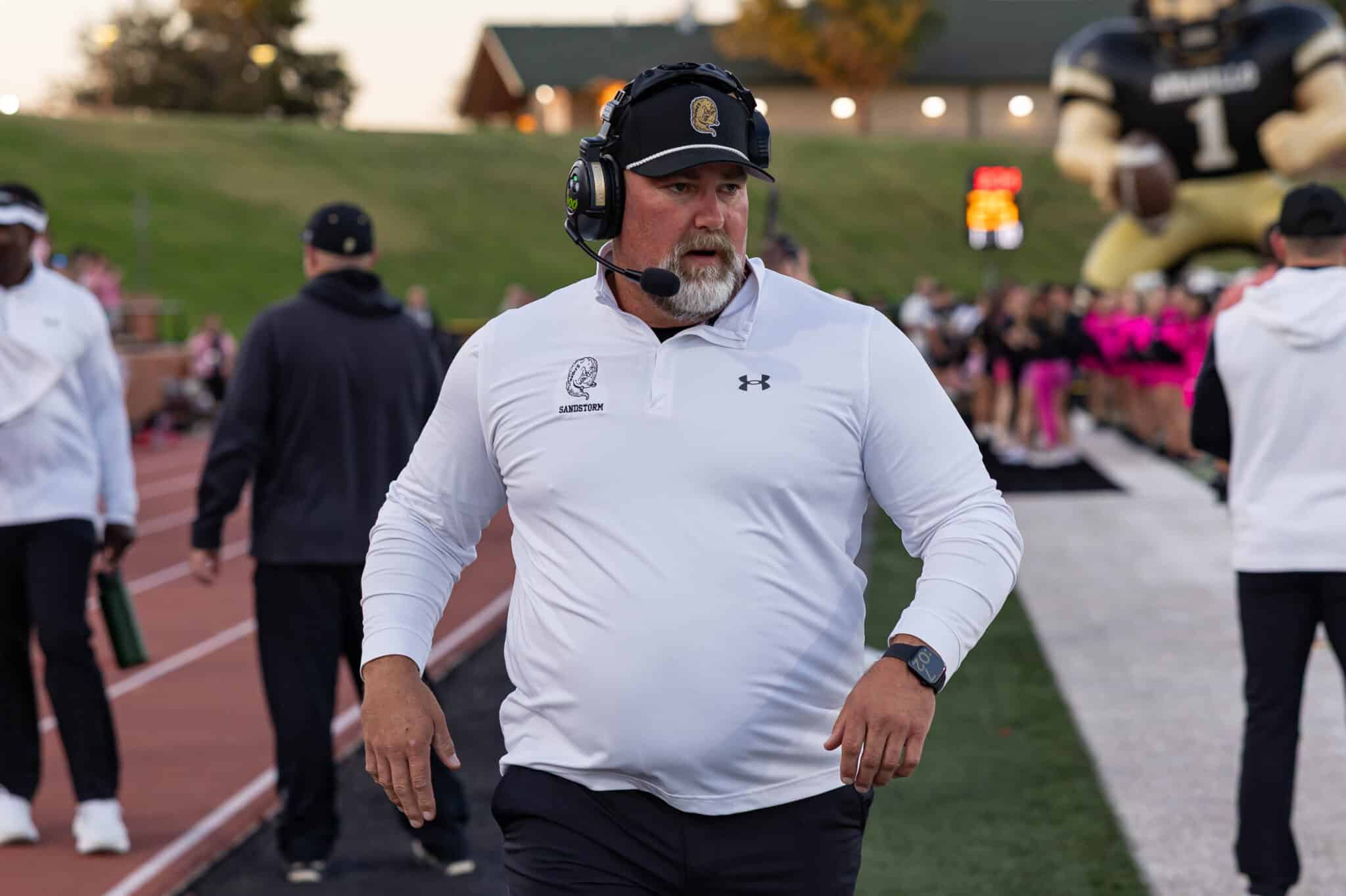 Male coach with a headset mic in a white polo on a football field, walking along the track with spectators in the background.]','Male coach wearing a headset and SandStorm polo, on the sideline during a game, crowd visible behind him.