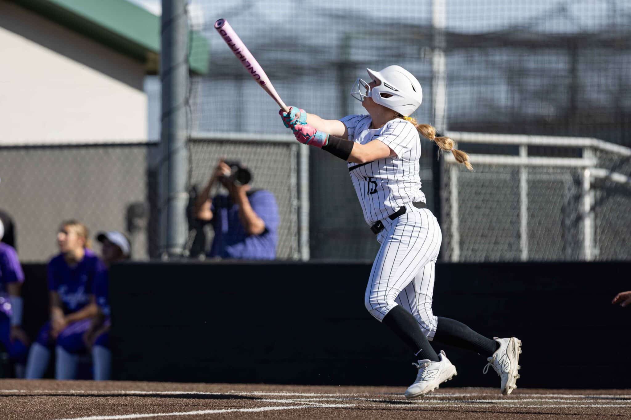Softball batter in a white pinstriped uniform and helmet swings a pink bat during a game.