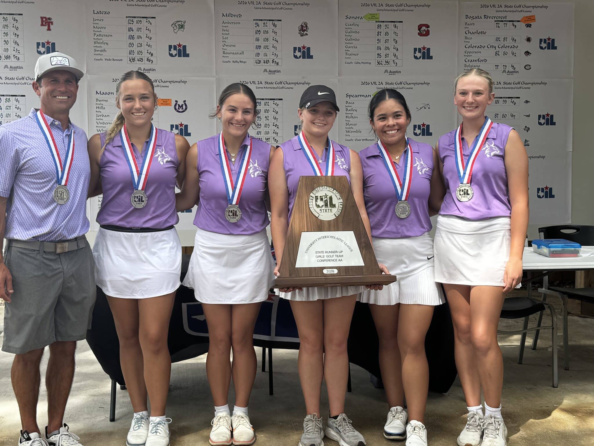 Coach and five young female golfers in purple shirts wearing medals, posing with a large award plaque.