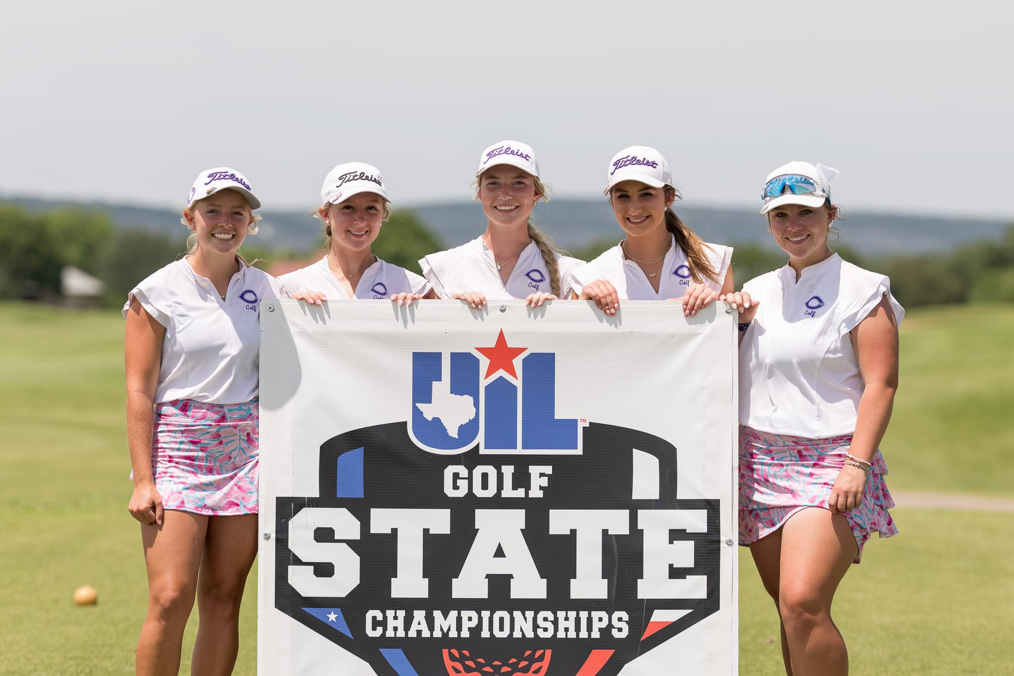 Five female golfers in white shirts and caps pose together behind a banner at a state golf championships event on a sunny course.