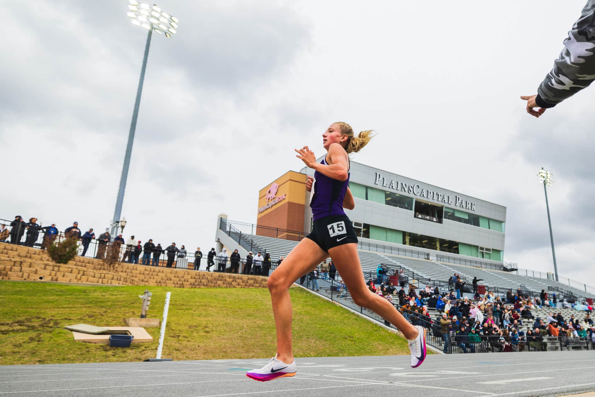 Female sprinter in a purple uniform runs on a track with a stadium and crowd behind.