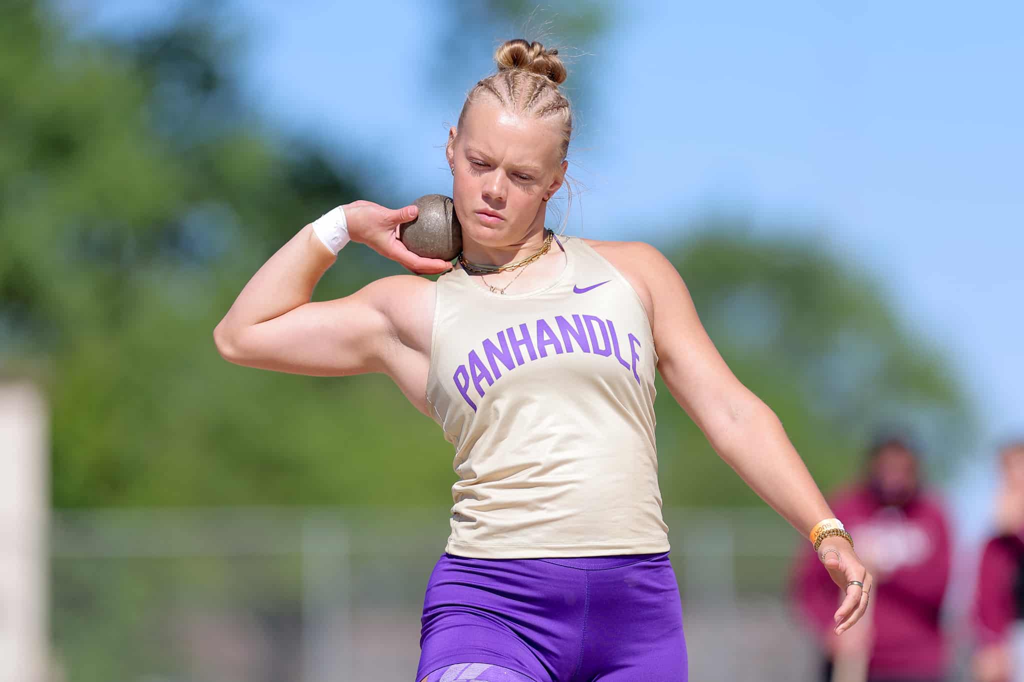Female track and field athlete in a beige singlet preparing to throw a shot put on a sunny day.