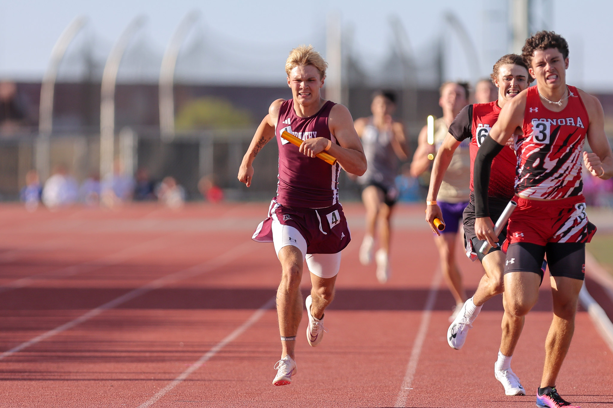 Male sprinters race on a red track, front runner in maroon holding a yellow baton as others chase behind.