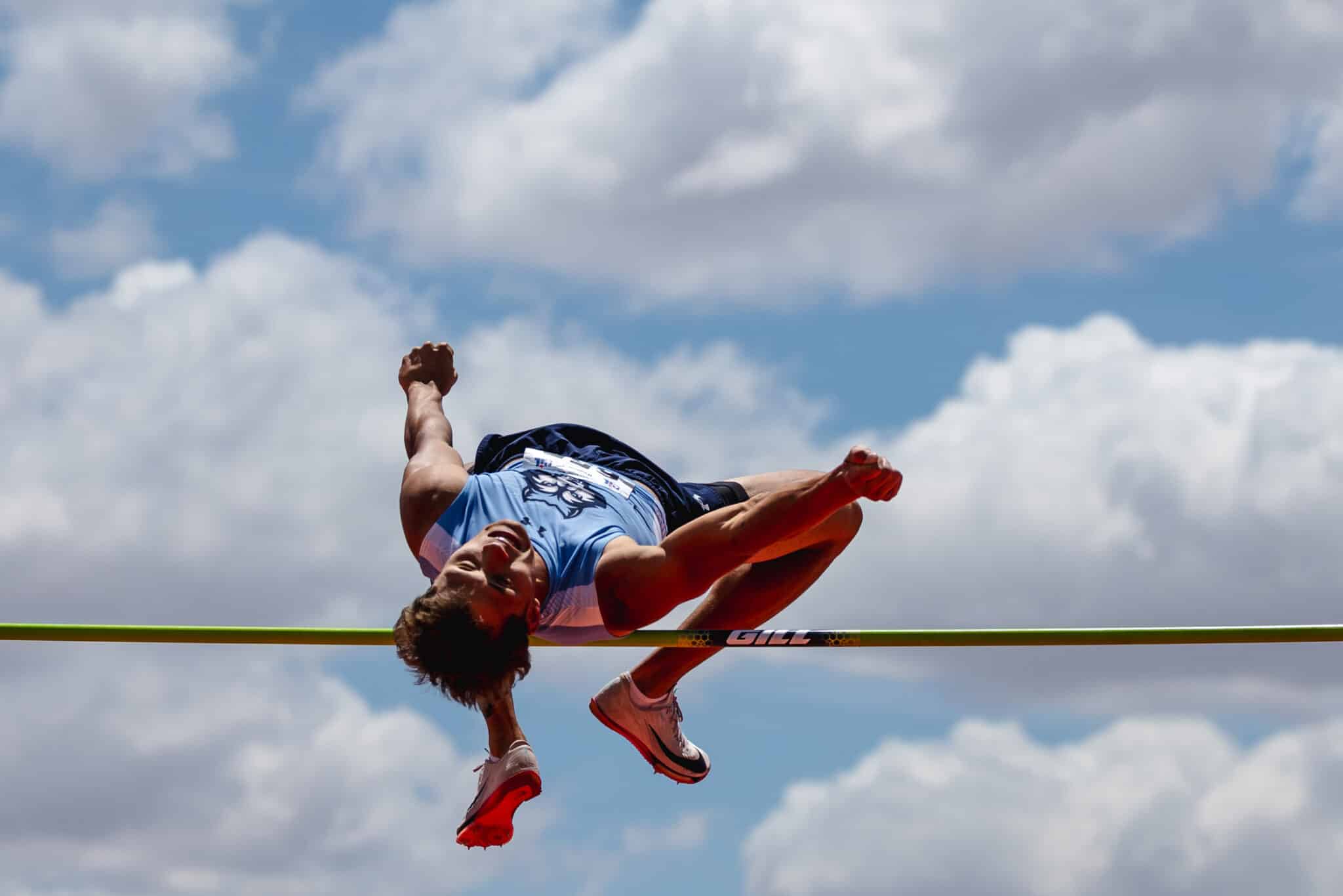 Athlete in a blue uniform clears a track hurdle midair against a blue sky with clouds.