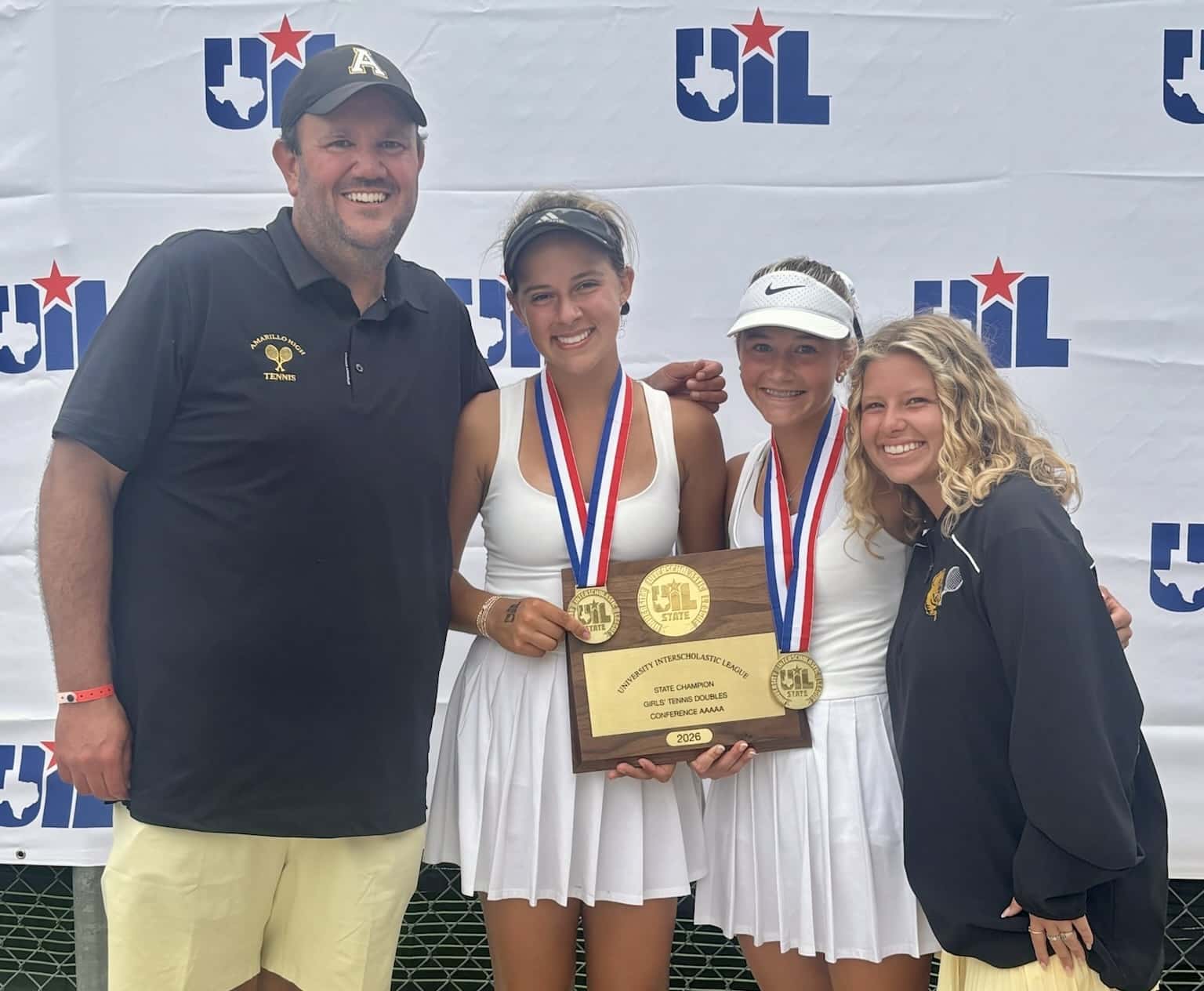 Two female tennis players in white dresses pose with medals on a wooden trophy, flanked by a man and a woman coach in front of a UIL backdrop.