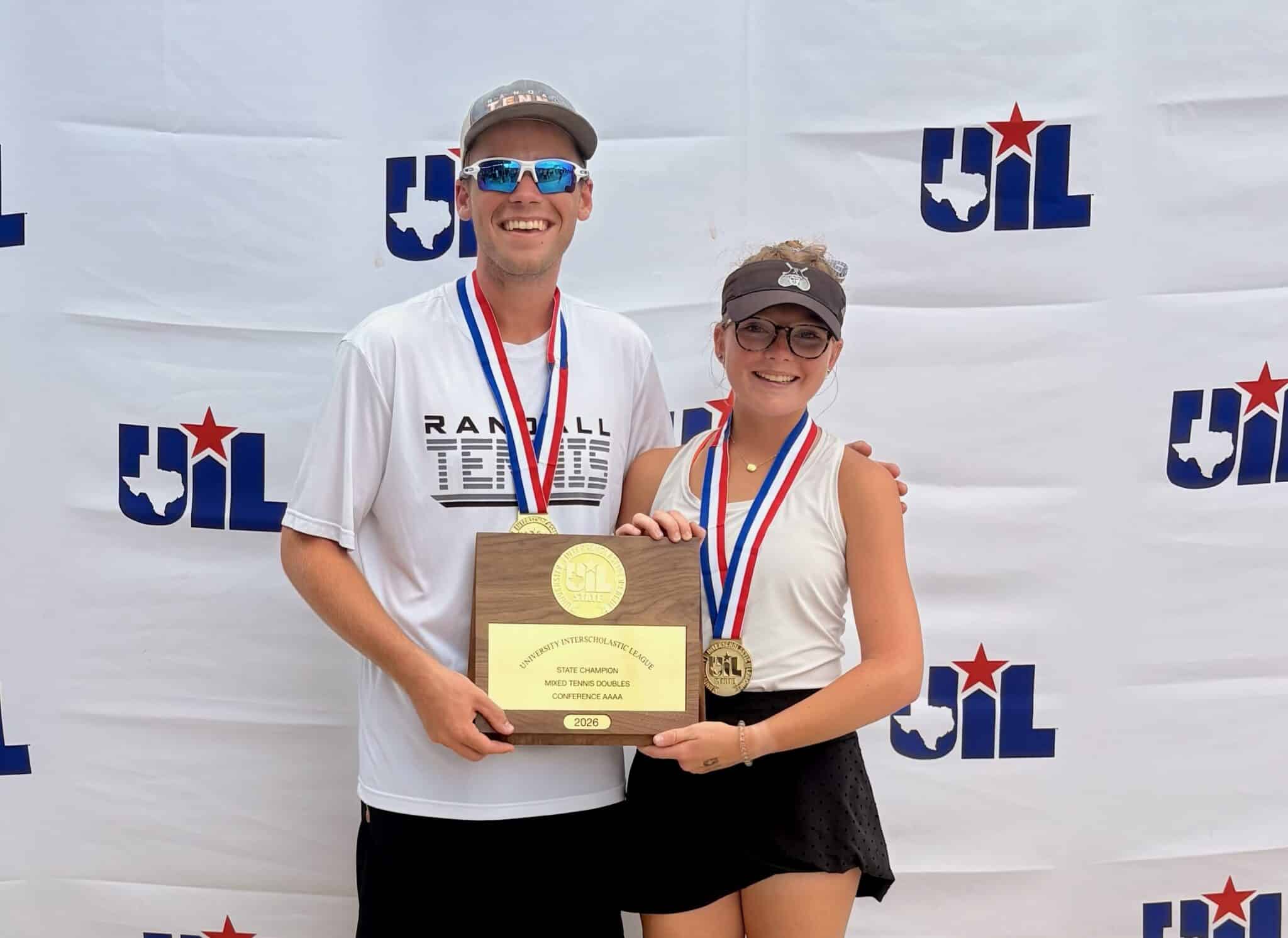Two young athletes pose with a wooden trophy and gold medals in front of a UIL backdrop with blue-red logos, smiling at the camera.