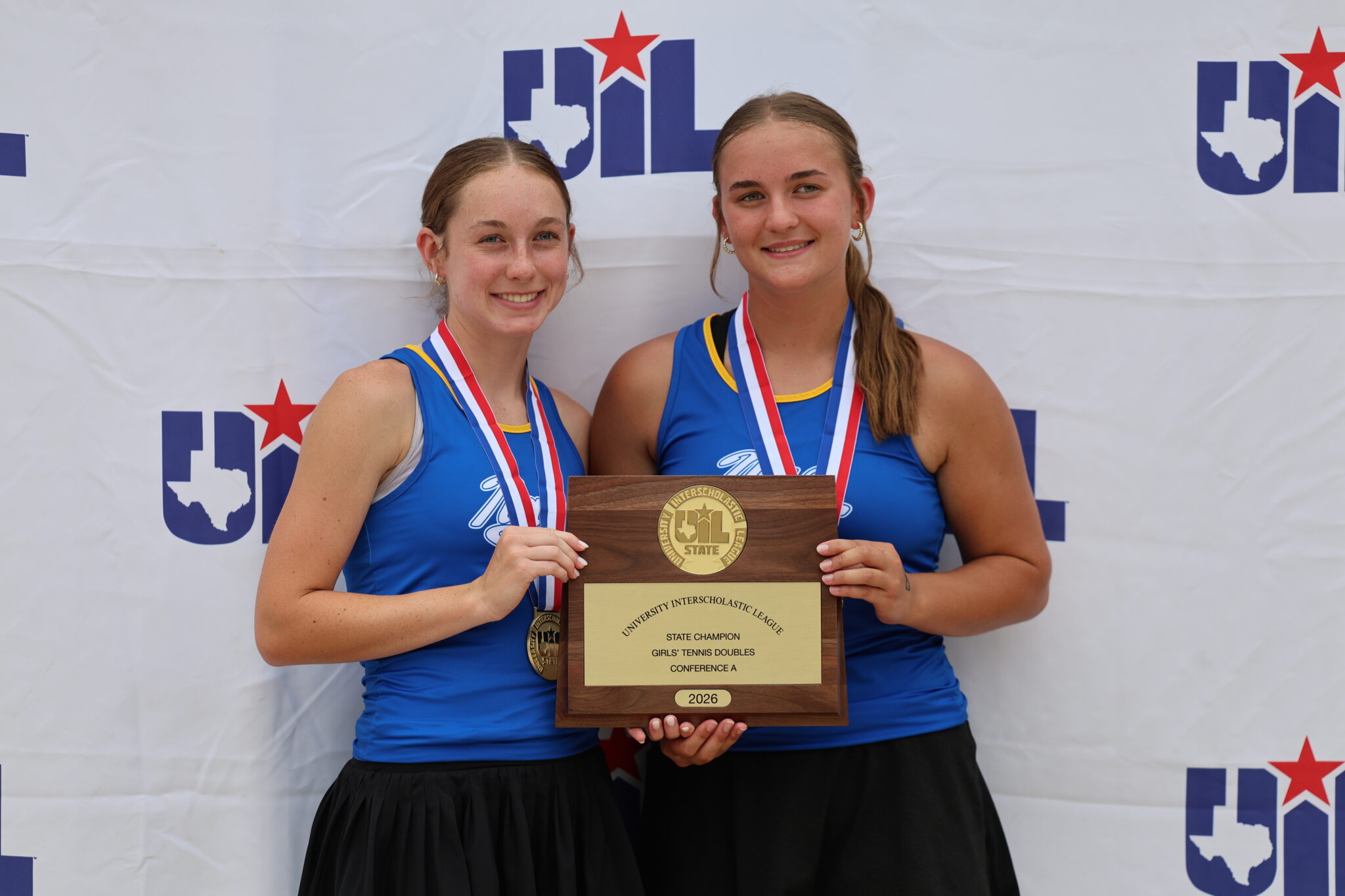 Two female athletes in blue uniforms smile while holding a large wooden trophy with a gold plaque and wearing medals around their necks, posing in front of a banner.