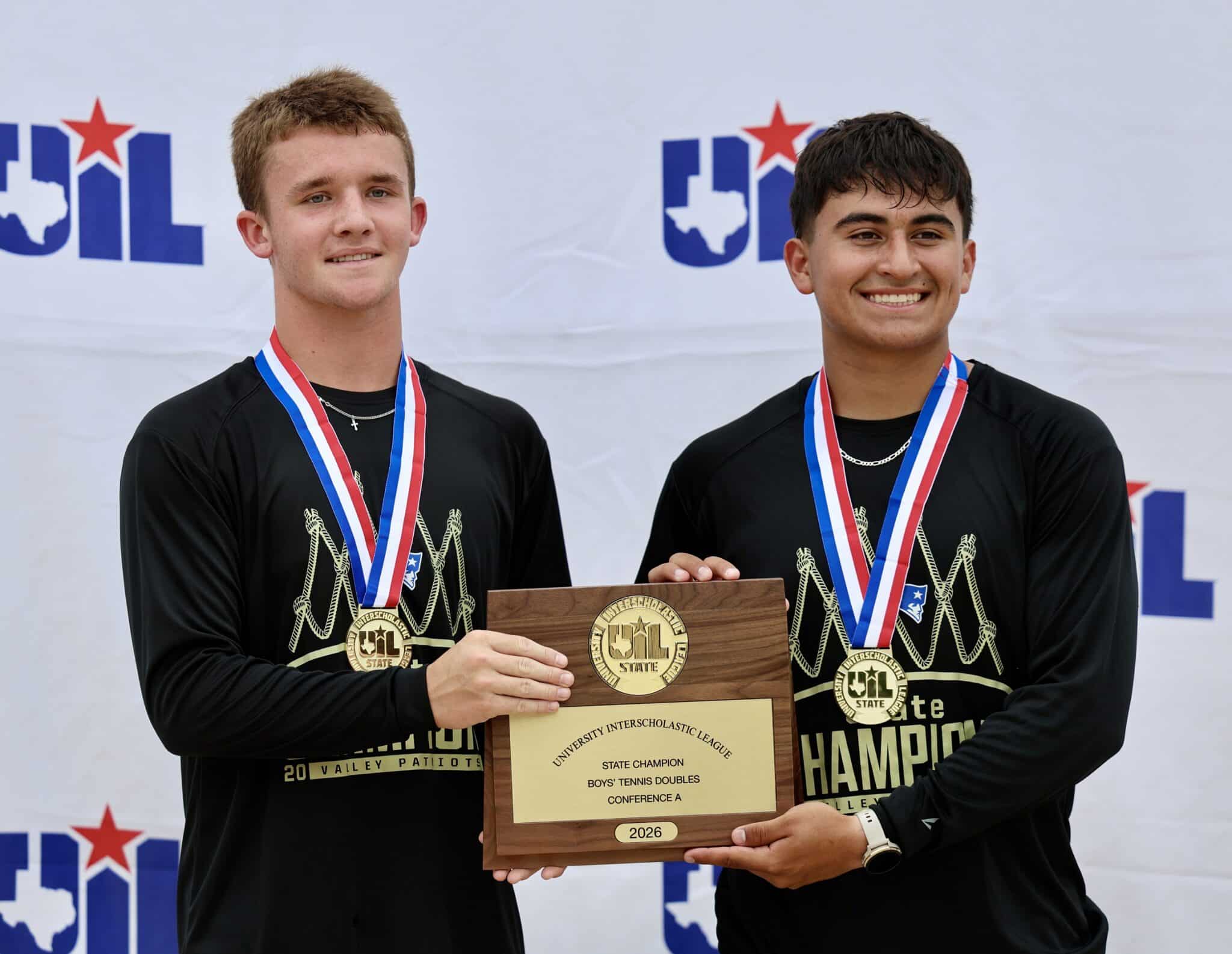 Two young athletes with medals pose beside a wooden plaque, indicating they are state champions in doubles tennis (2026).