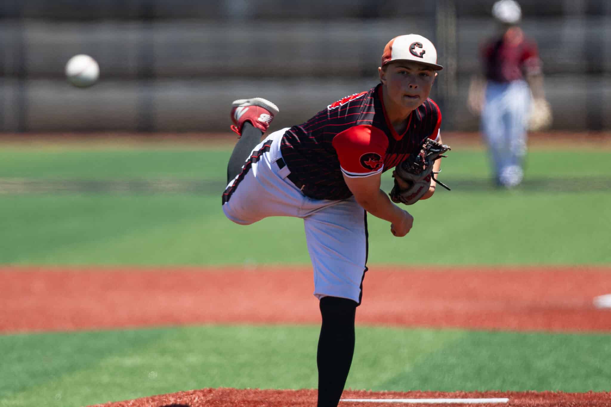 Baseball pitcher in red and black uniform delivering a pitch on the mound, ball in midair nearby.