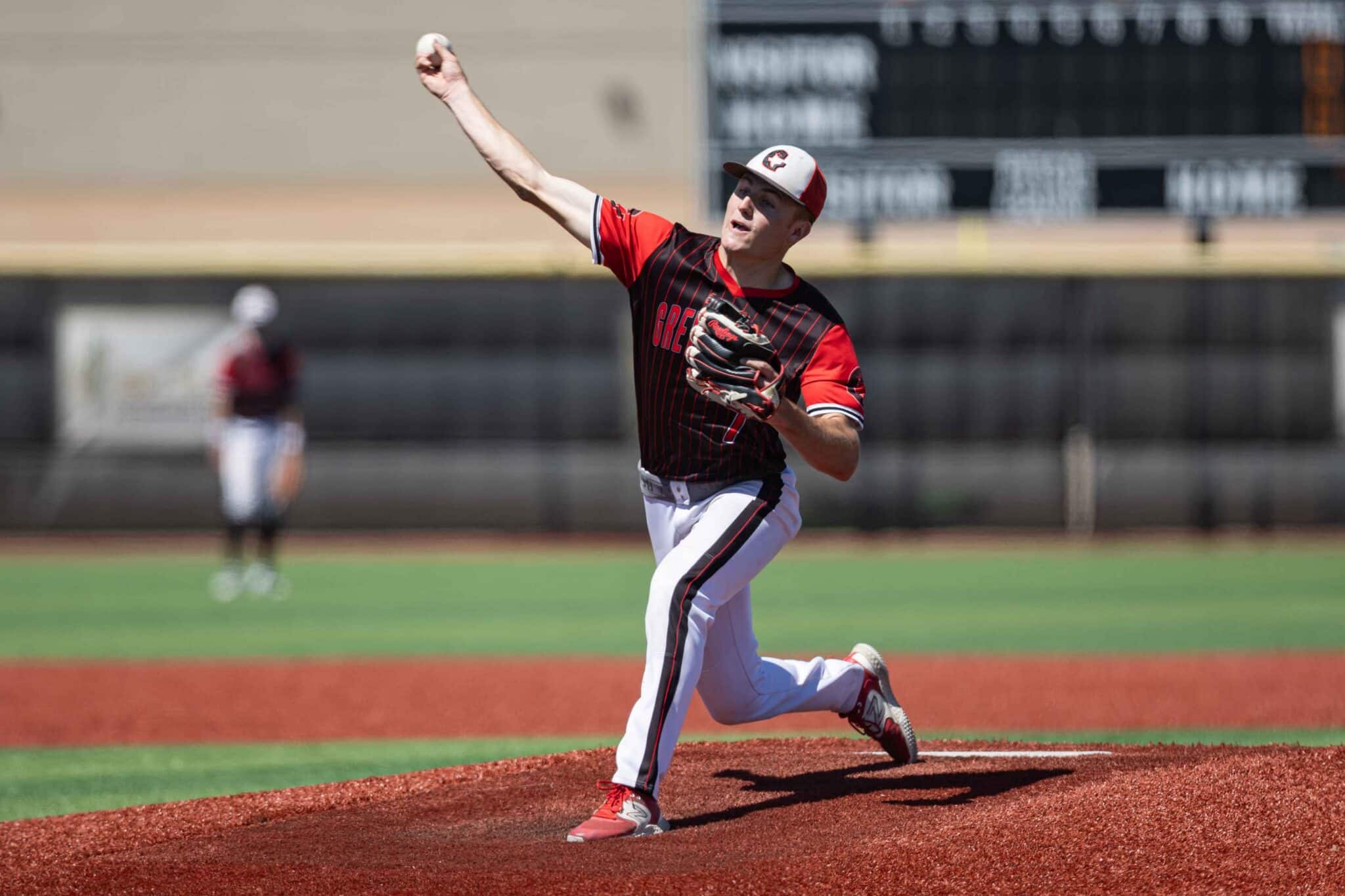 Male baseball pitcher in red and black uniform on the mound, mid-throw with the ball raised in his right hand.