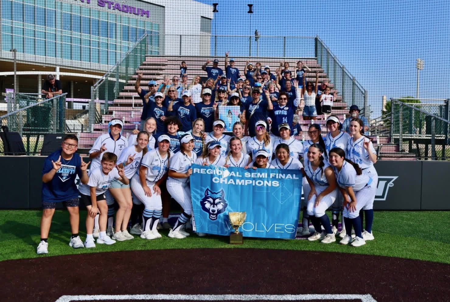 Girls softball team poses on the field holding a 'Regional Semifinal Champions' banner with a trophy in front of bleachers and fans in blue.