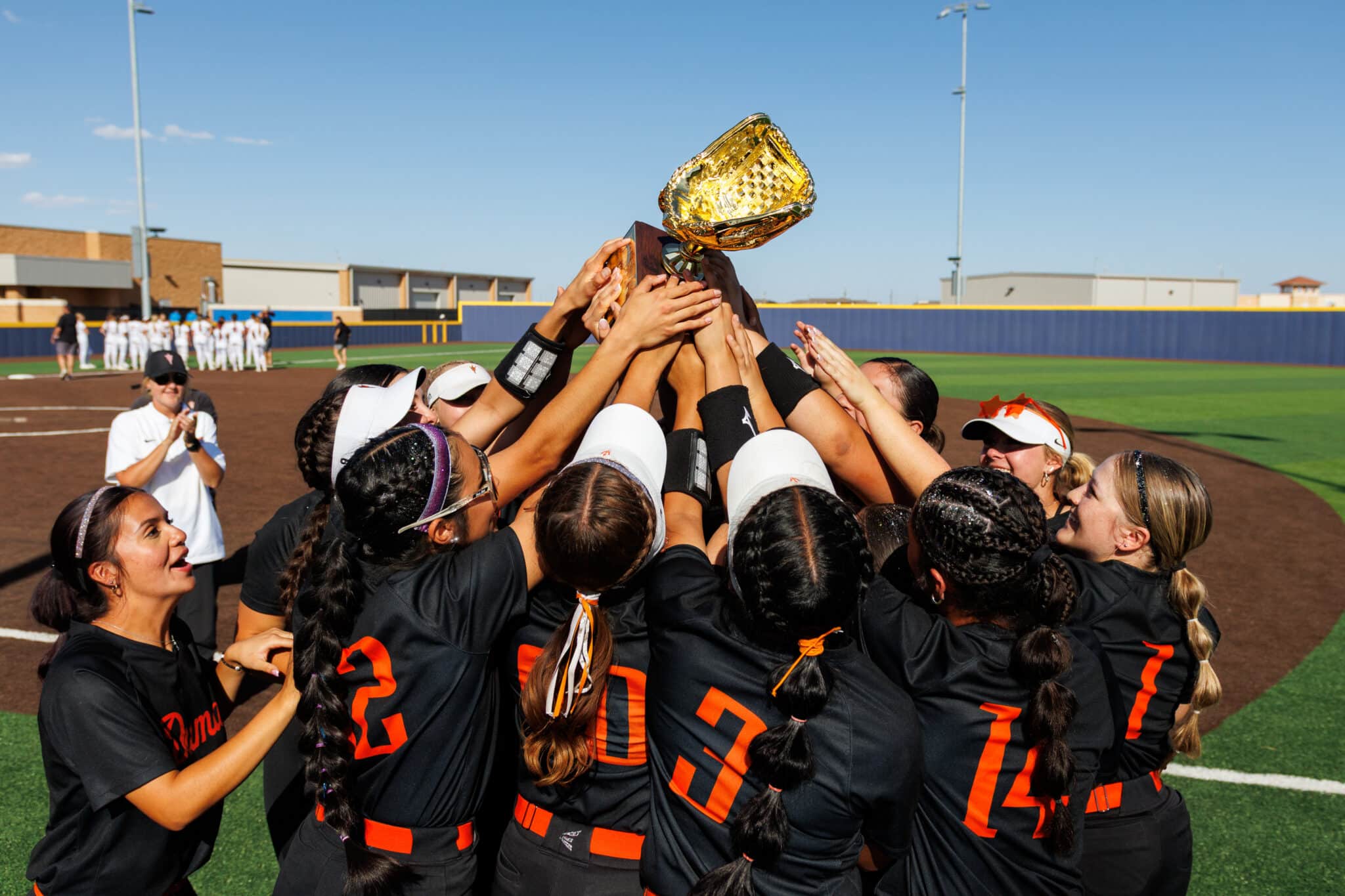 IMG_3221JPEG | Press Pass Sports A group of athletes in black jerseys huddles and raises a large golden trophy on a baseball field during a sunny day.