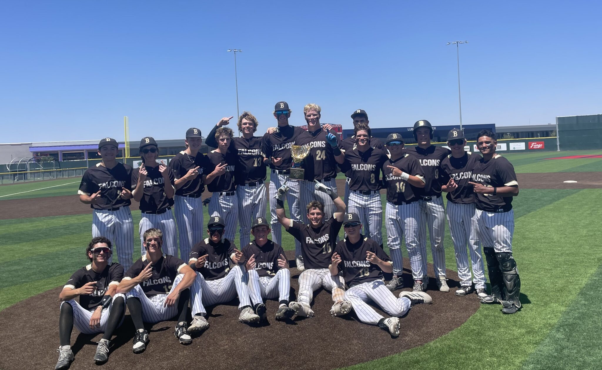 Falcons baseball team posing on the infield with a trophy after a win, players in black uniforms with white pinstripe pants.